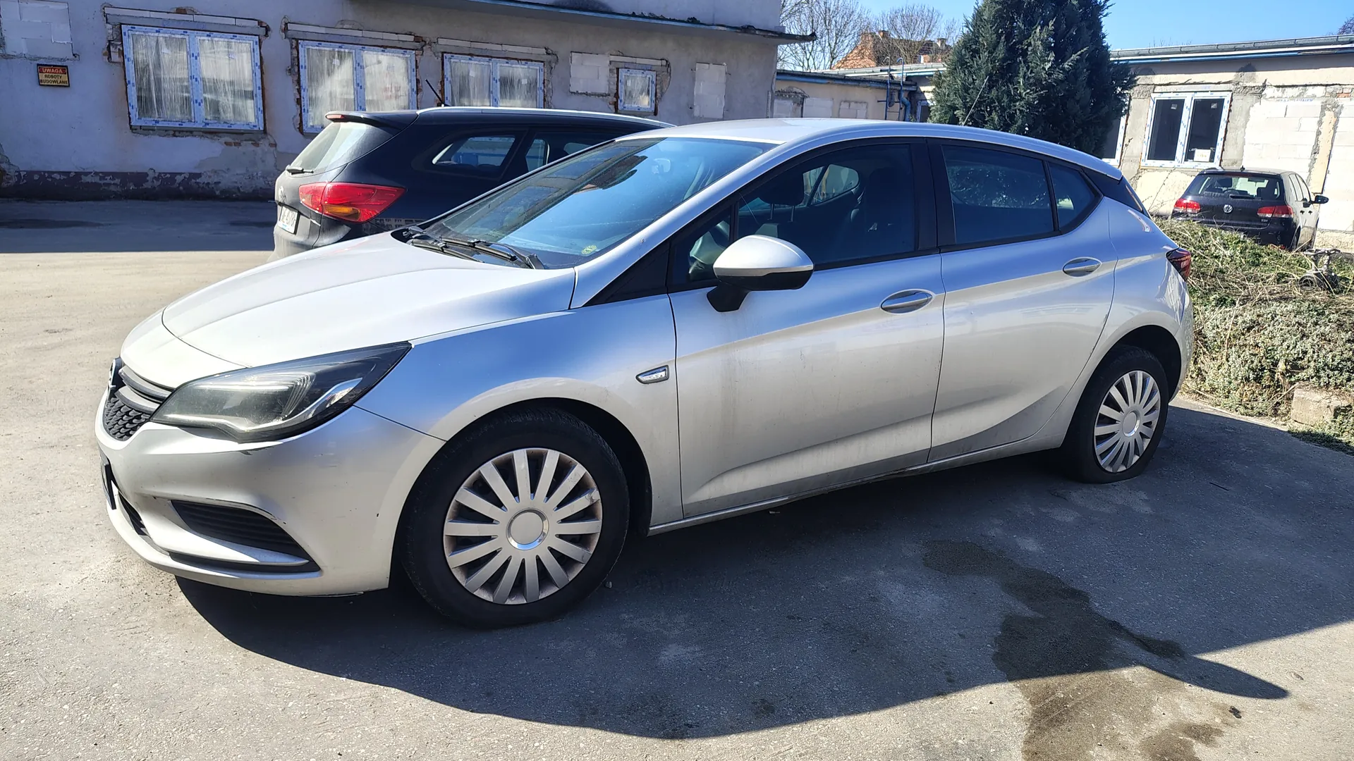 Srebrny samochód typu hatchback zaparkowany na betonowym placu. W tle widoczne są dwa inne samochody: czarny i ciemnoszary. Za nimi stoją budynki z oknami zasłoniętymi zasłonami oraz fragmenty ścian z widocznymi uszkodzeniami. Po prawej stronie obrazu znajduje się krzak. Na ścianie budynku po lewej stronie widoczna jest tabliczka z napisem "UWAGA". Słoneczna pogoda, cienie wskazują na jasne światło dzienne.