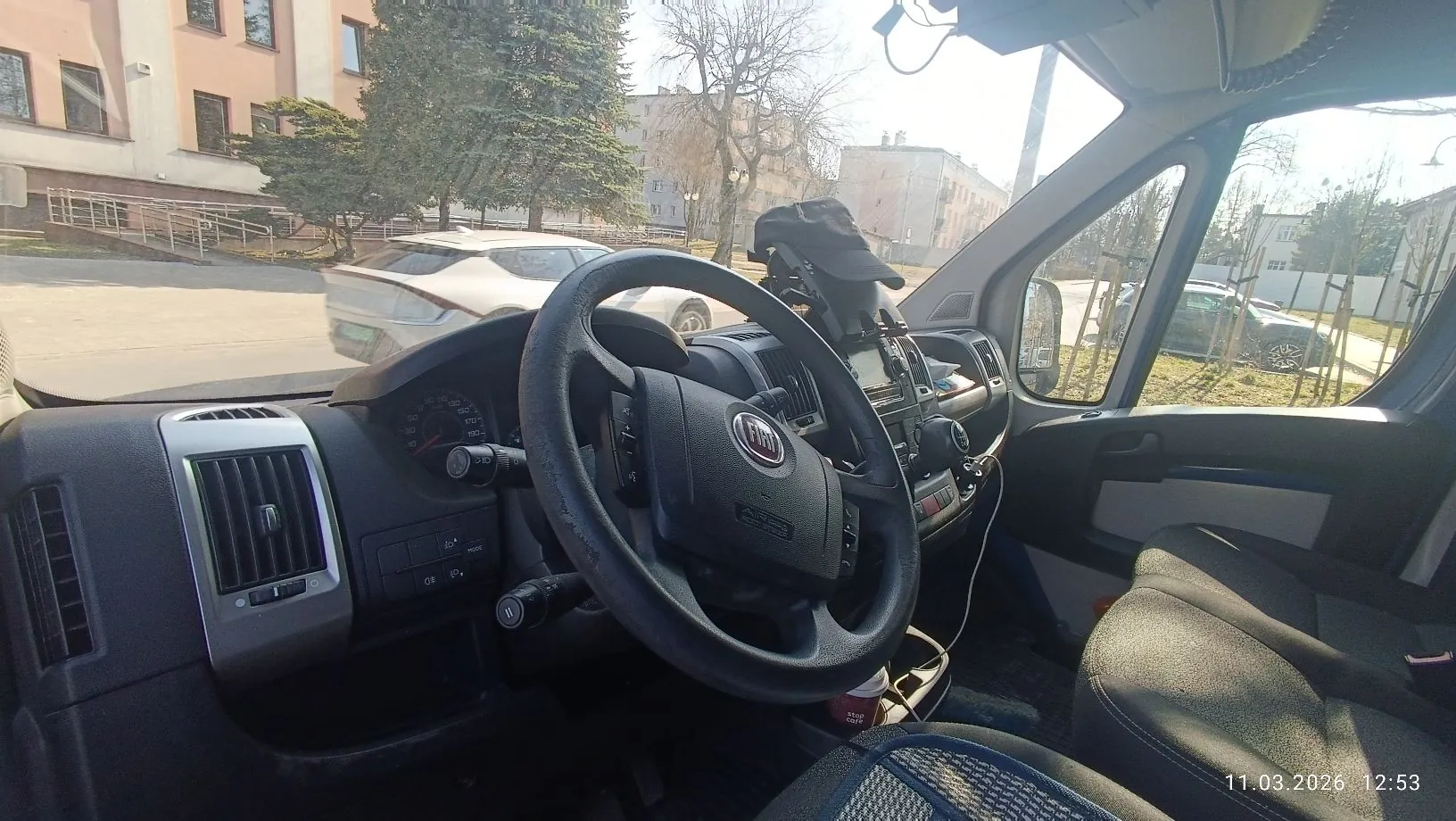 Widok z wnętrza samochodu marki Fiat, z perspektywy kierowcy. Widoczna jest czarna kierownica z logo Fiat, deska rozdzielcza, wskaźniki prędkości oraz przednia szyba. Przez szybę widać zaparkowany biały samochód, budynek mieszkalny oraz drzewa. W prawym dolnym rogu znajduje się data i godzina: 11.03.2026 12:53.