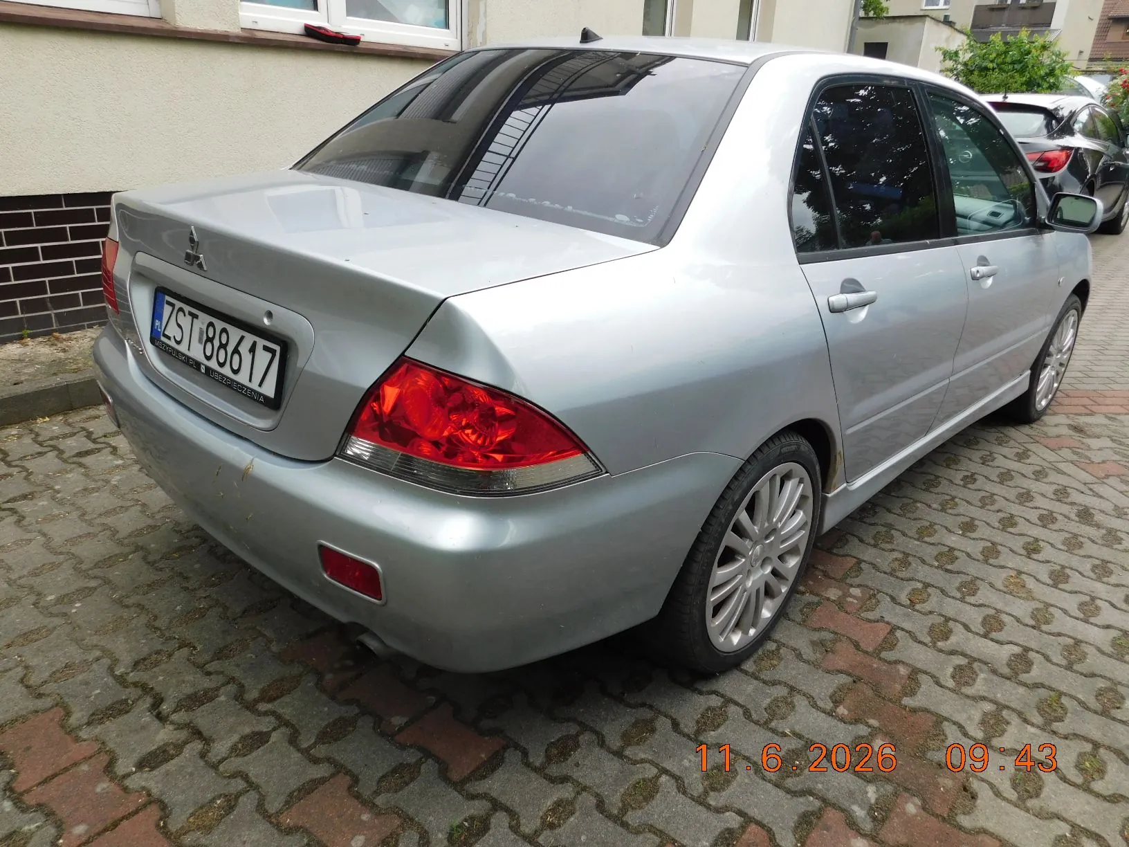 Srebrny samochód osobowy marki Mitsubishi w nadwoziu sedan, zaparkowany tyłem na kostce brukowej. Widoczna jest tablica rejestracyjna o numerze ZST 88617. W prawym dolnym rogu zdjęcia znajduje się pomarańczowy znacznik daty i godziny: 11.6.2026 09:43.