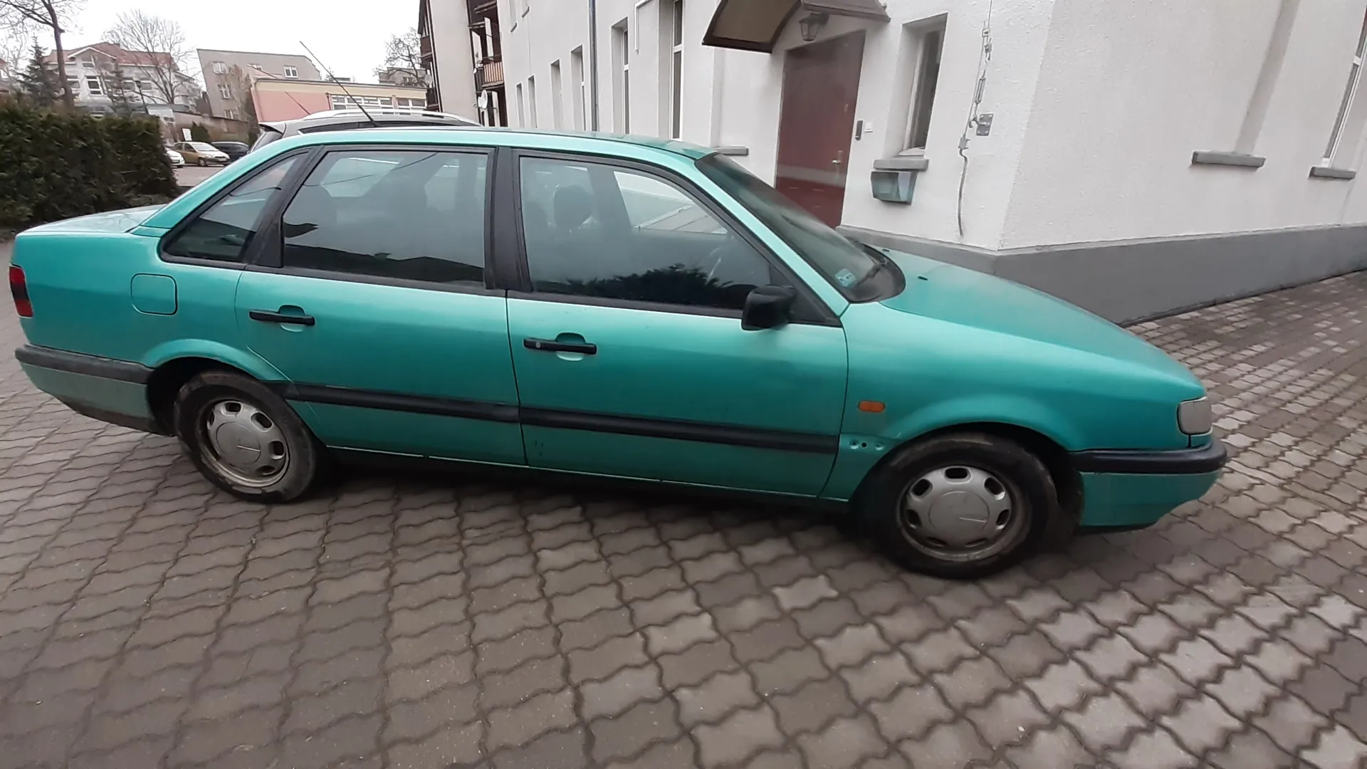 Zdjęcie przedstawia turkusowy samochód osobowy typu sedan zaparkowany na brukowanej powierzchni. Pojazd ma czarne listwy boczne, srebrne felgi i ciemne szyby. W tle widoczny jest biały budynek z brązowymi drzwiami, oknami oraz szarą podstawą. Za samochodem znajdują się krzewy, inne budynki mieszkalne oraz częściowo widoczny inny pojazd. Na tle niebo jest pochmurne.