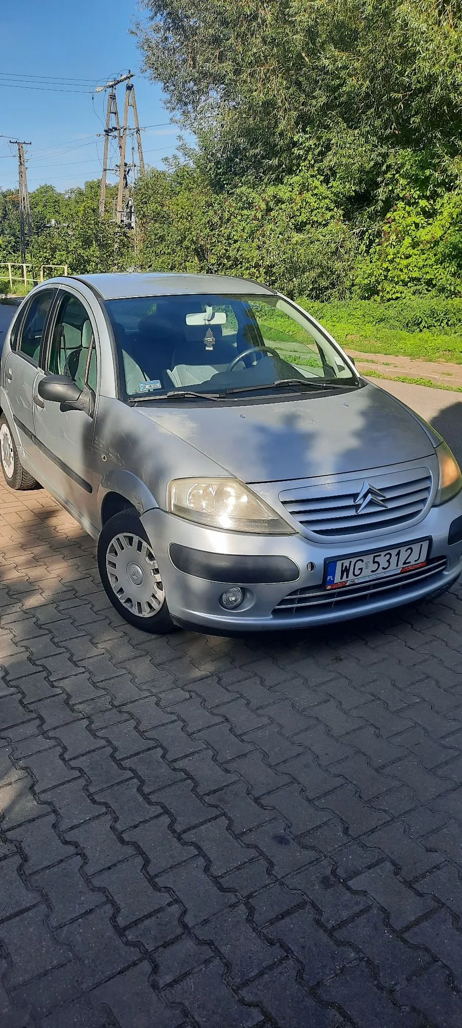 Srebrny samochód osobowy marki Citroën zaparkowany na kostce brukowej. Pojazd ma polską tablicę rejestracyjną o numerze WG 5312J. W tle widoczne są zielone drzewa, krzewy oraz słupy linii energetycznej na tle niebieskiego nieba.