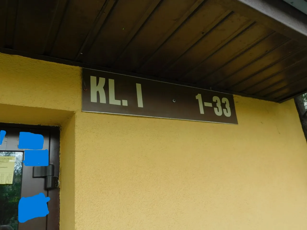 Zdjęcie fragmentu żółtej ściany budynku pod brązowym podbitkiem dachu. Na ścianie zamontowana jest prostokątna, ciemna tabliczka z białym napisem „KL. I 1-33”. Po lewej stronie widoczny jest fragment brązowych drzwi z niebieskimi zakryciami na szybach.