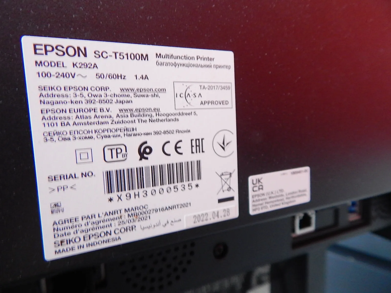 Zbliżenie na białą tabliczkę znamionową drukarki EPSON SC-T5100M. Na etykiecie widnieją informacje: model K292A, napięcie 100-240V~ 50/60Hz 1.4A, adresy producenta SEIKO EPSON CORP. w Japonii oraz EPSON EUROPE B.V. w Holandii. Tekst pojawia się również w języku cyrylicy. Widoczne są certyfikaty CE, EAC oraz logo ICASA APPROVED. Na dole znajduje się kod kreskowy z numerem seryjnym * X 9 H 3 0 0 0 5 3 5 *, data 2022.04.28 oraz napis MADE IN INDONESIA.