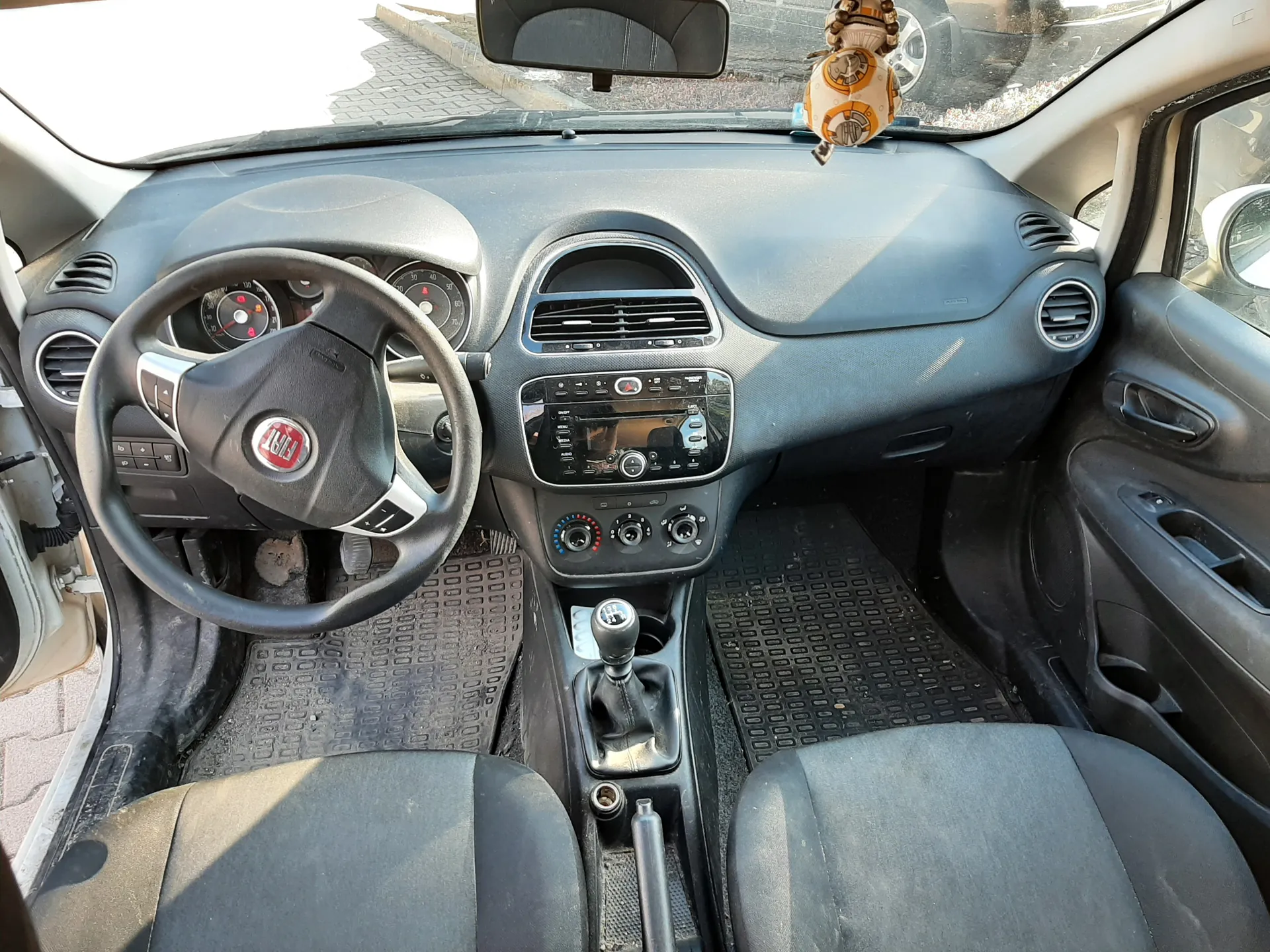 Wnętrze samochodu Fiat z widokiem na deskę rozdzielczą, kierownicę z logo Fiat, centralną konsolę z odtwarzaczem i przyciskami klimatyzacji, dźwignią zmiany biegów oraz czarnymi dywanikami podłogowymi. Na lusterku wstecznym wisząca zabawka w kształcie BB-8. Widoczne są również przednie siedzenia, otwarte drzwi po lewej stronie oraz fragment zewnętrzny przez przednią szybę z chodnikiem i innym pojazdem.