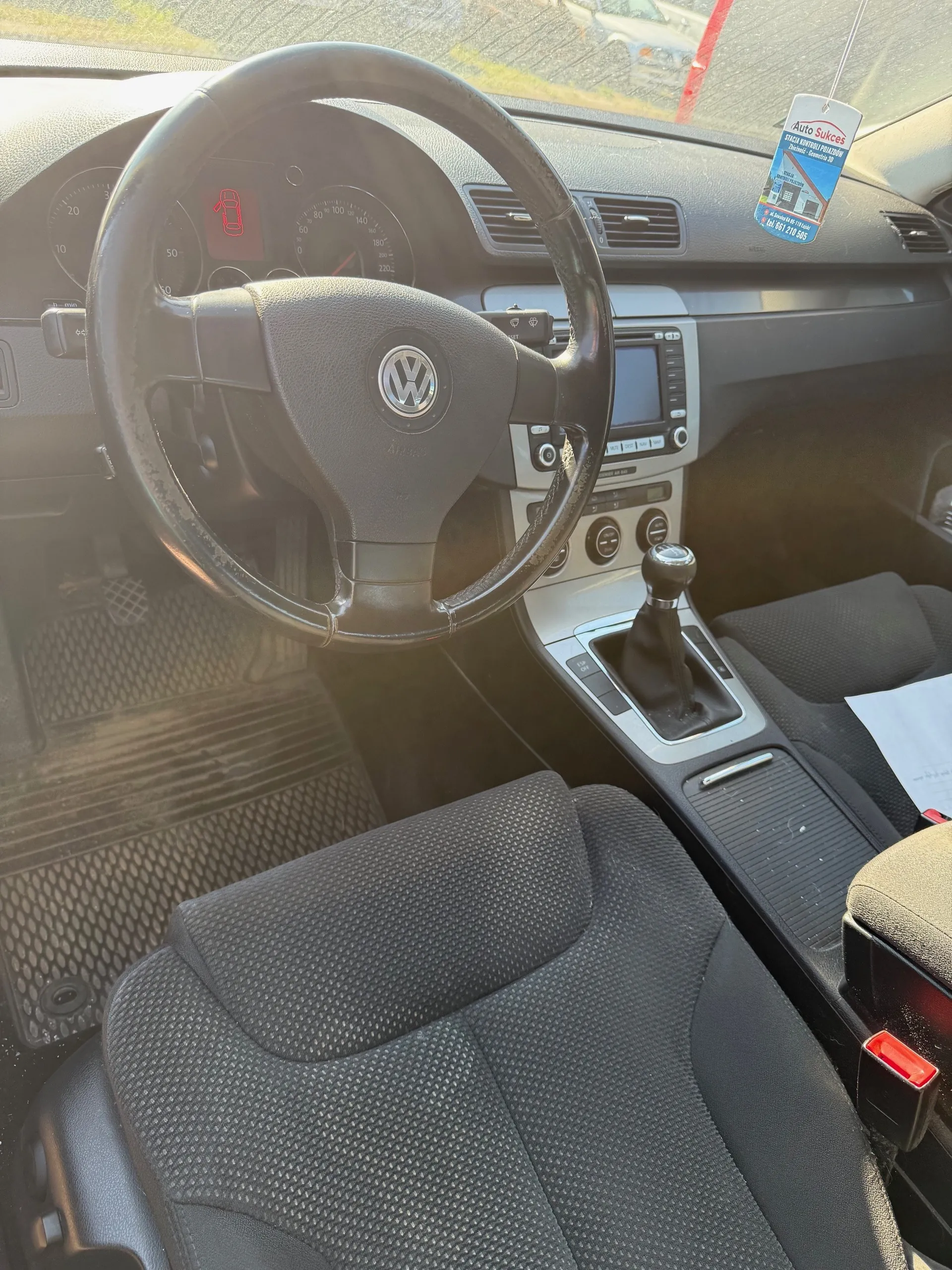 Wnętrze samochodu marki Volkswagen z widocznym kierownicą z logo VW, tablicą rozdzielczą z prędkościomierzem i czerwonym sygnalizatorem w kształcie samochodu, centralną konsolą z ekranem i dźwignią zmiany biegów, czarnymi siedzeniami tapicerowanymi tkaniną oraz dywanikami podłogowymi. Na lusterku wstecznym wiszący znaczek z napisem 'Auto Sukces'.