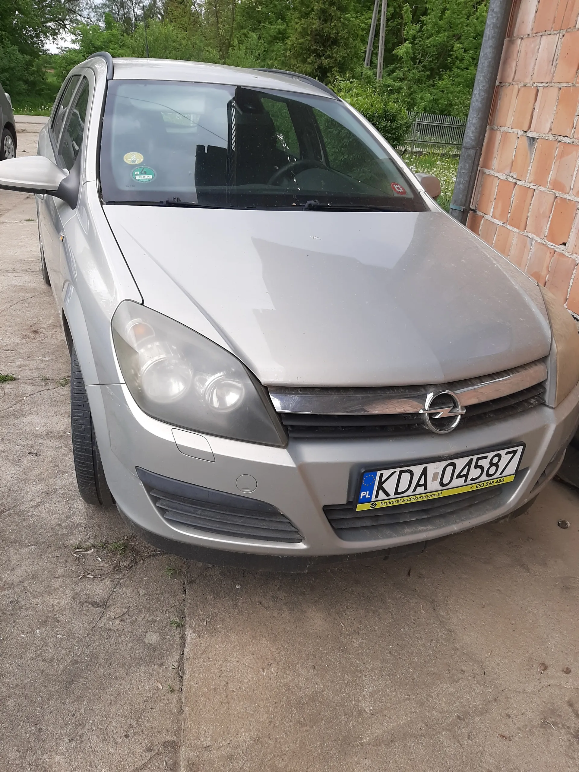 Srebrny samochód z tablicą rejestracyjną KDA 04587. W tle ceglana ściana, zielone drzewa i betonowa powierzchnia. Na przedniej szybie znajdują się naklejki.