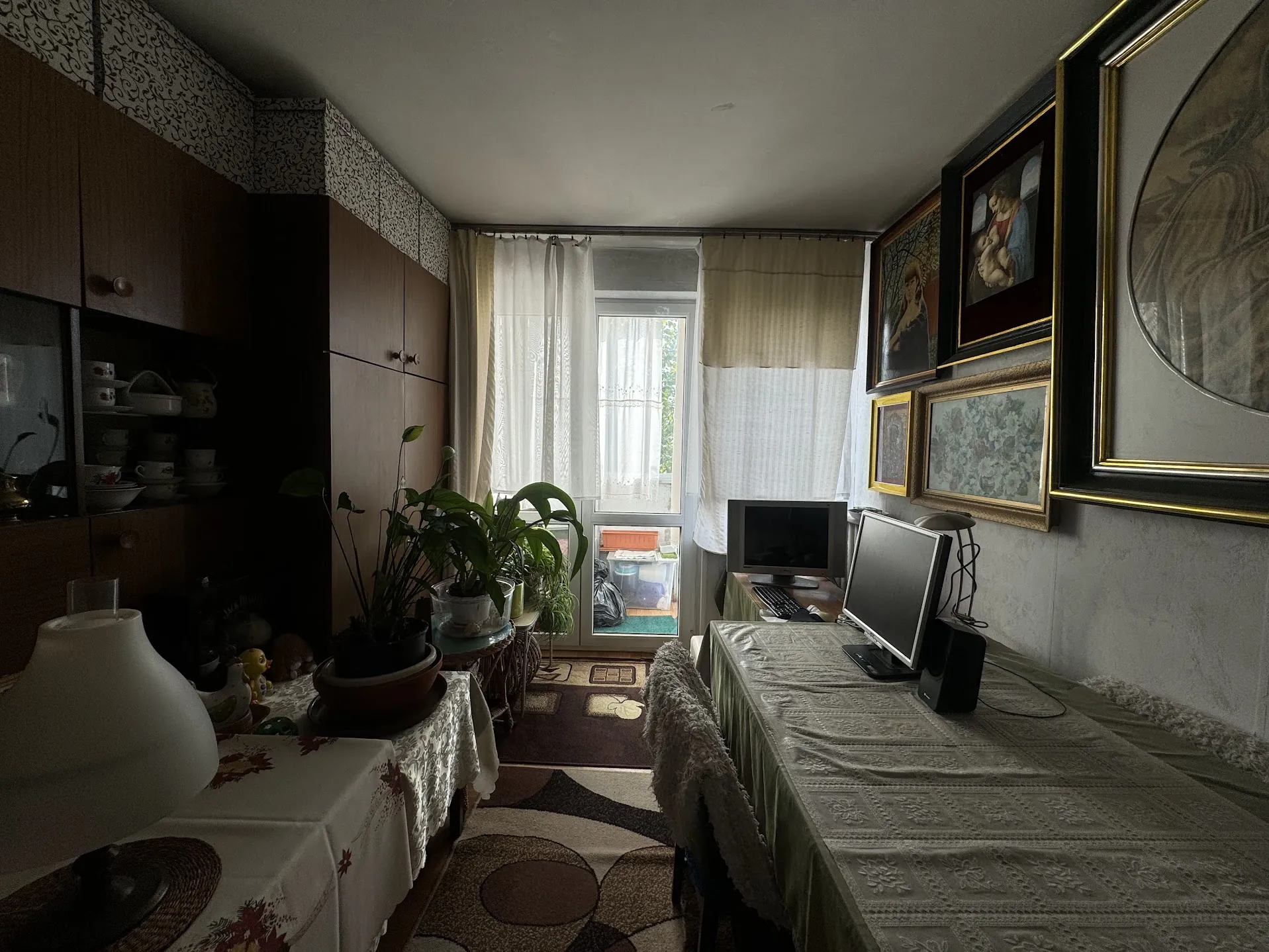 Wnętrze pomieszczenia z meblami i dekoracjami. Po lewej stronie drewniana szafka z szklanymi drzwiami zawierająca porcelanowe naczynia. Na stole z białym obrusem z czerwonymi wzorami znajdują się doniczki z roślinami, lampa oraz małe przedmioty, w tym żółta kaczka. W centrum pomieszczenia okno z białymi zasłonami, za którym widoczny jest balkon z przedmiotami. Po prawej stronie stół z zielonym obrusem, na którym stoją dwa monitory komputerowe, klawiatura i głośnik. Ściany zdobione ramkami z obrazami i fotografiami. Podłoga wyłożona dywanem z geometrycznym wzorem.