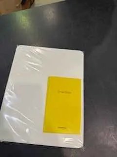 Biały prostokątny przedmiot owinięty przezroczystą folią, z żółtym prostokątnym etykietą na górze. Tło stanowi ciemna powierzchnia, prawdopodobnie stół. Na folii widoczne są odbicia światła. Tekst na żółtej etykiecie nieczytelny.