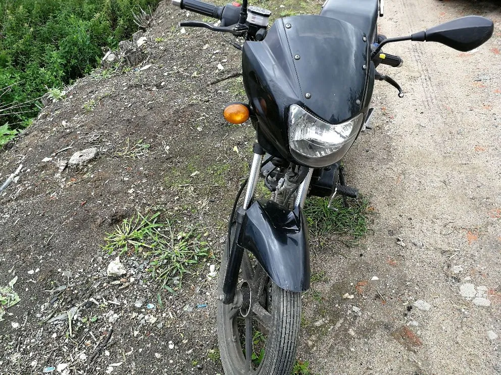 Zbliżenie na przód czarnego motocykla zaparkowanego na piaszczystej drodze. Widoczna jest przednia lampa, owiewka, kierownica z lusterkiem oraz przednie koło z błotnikiem. Po lewej stronie znajduje się nasyp z ziemi i zielona roślinność.