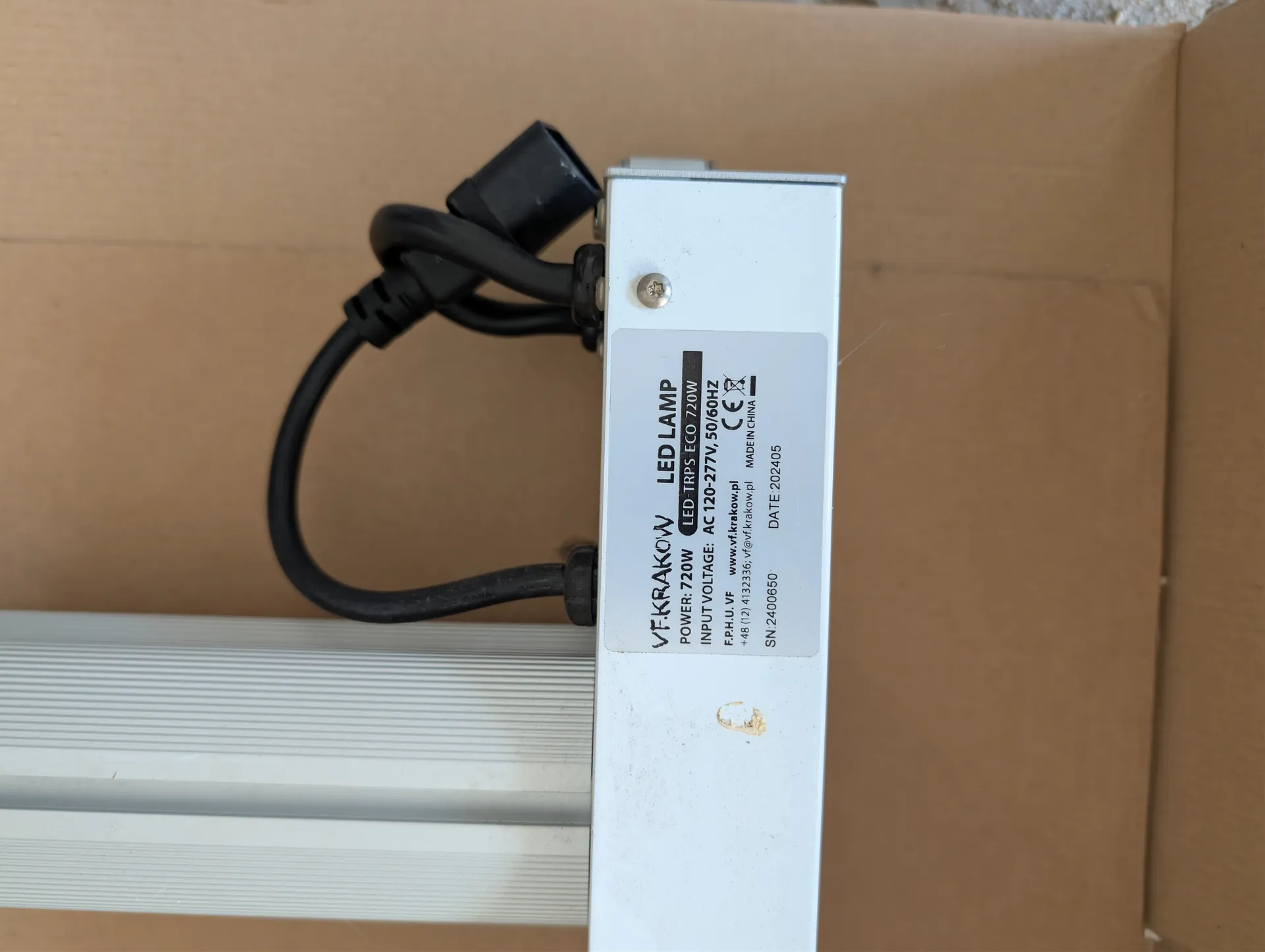 Biała lampa LED z białą etykietą zawierającą tekst: VF.KRAKOW LED LAMP, POWER: 720W, LED-TRPS ECO 720W, INPUT VOLTAGE: AC 120-277V, 50/60HZ, F.P.H.U. VF, www.vf.krakow.pl, +48 (12) 4132336; vf@vf.krakow.pl, MADE IN CHINA, SN:2400650, DATE:202405. Czarny przewód zasilający zwinięty na górze lampy. Tło z brązowego kartonu. Na białej powierzchni lampy widoczne jest małe uszkodzenie.
