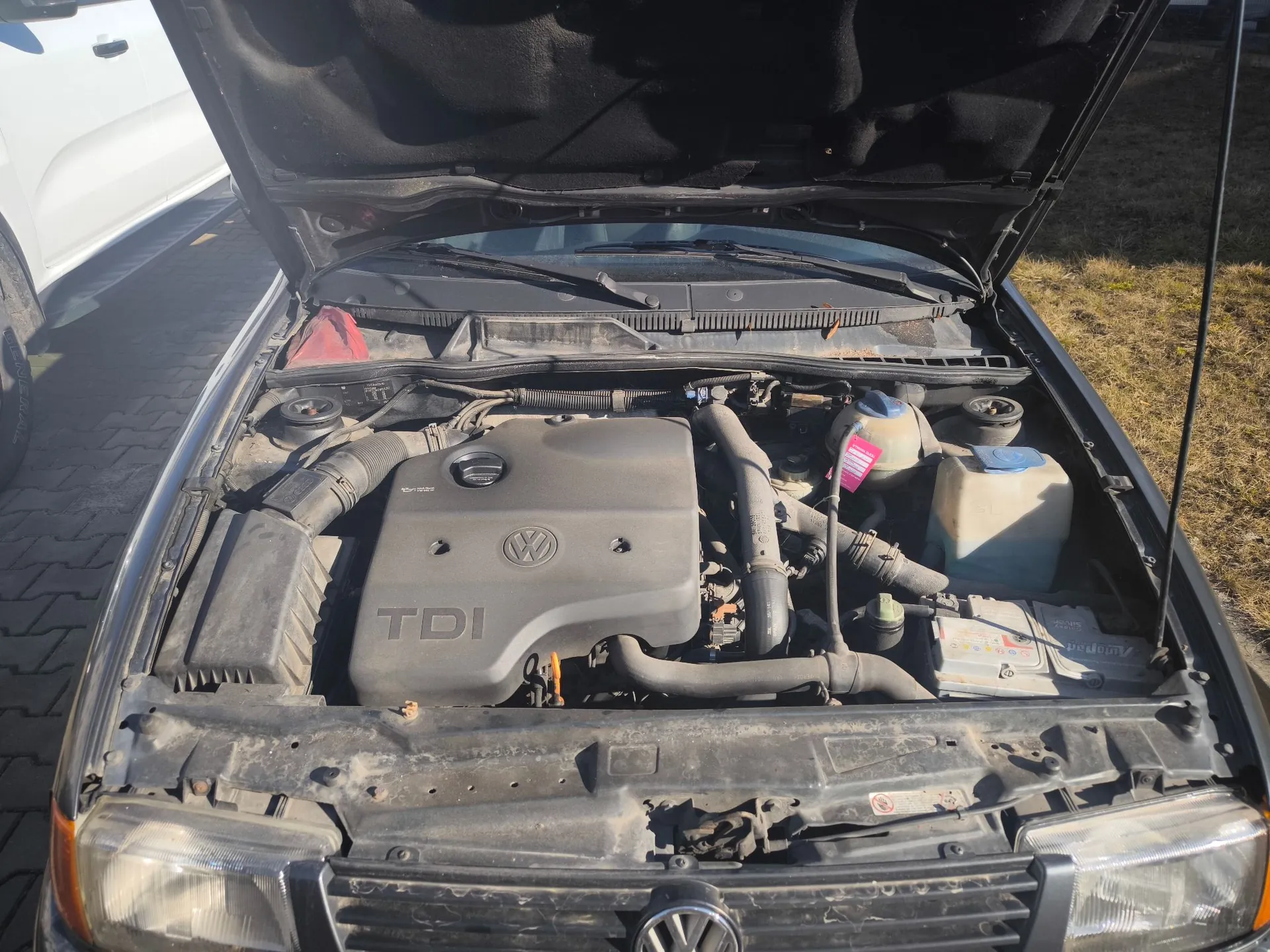 Widok z góry na otwartą maskę samochodu, ukazującą silnik diesla. Na plastikowej osłonie silnika widoczne jest logo Volkswagena oraz napis 'TDI'. W komorze silnika znajdują się różne przewody, zbiorniki z płynami oraz akumulator. Samochód jest w kolorze ciemnym, a w tle widoczny jest fragment białego pojazdu oraz kostka brukowa.
