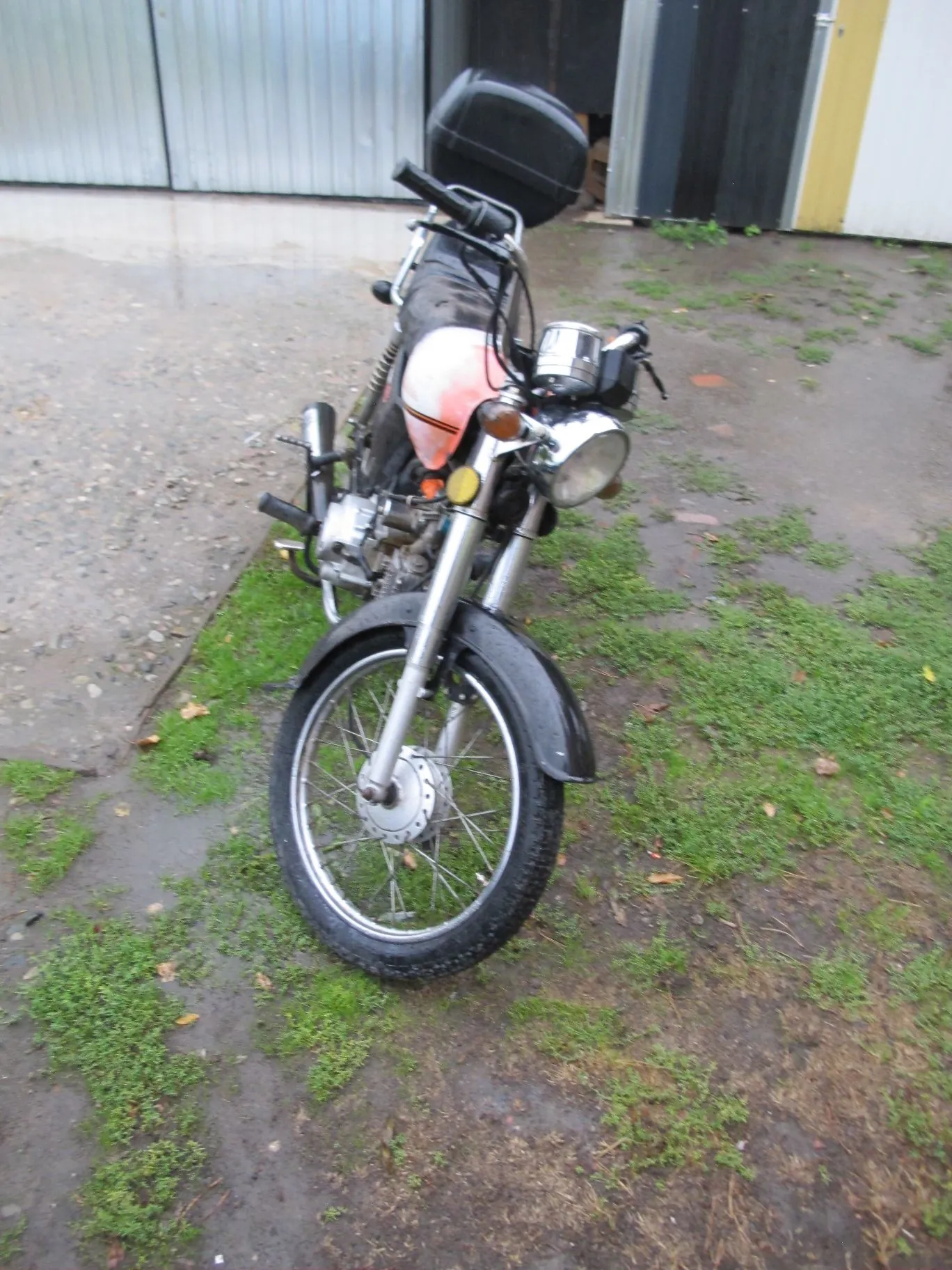 Motocykl zaparkowany na mokrym, częściowo trawiastym podłożu przed budynkiem z blachy falistej. Pojazd ma srebrny przedni widelec, szprychowe koła, okrągły reflektor oraz czerwony zbiornik paliwa. Z tyłu znajduje się czarny kufer.