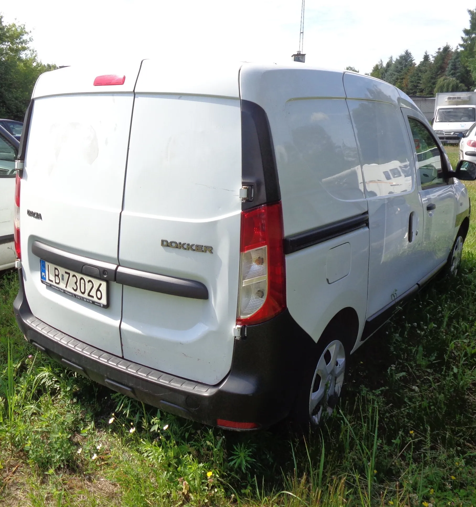 Biały samochód dostawczy marki Dacia model Dokker, zaparkowany na trawie. Widoczny jest tył i prawy bok pojazdu. Samochód posiada tablicę rejestracyjną o numerze LB 73026. Na klapie bagażnika znajduje się napis DOKKER.