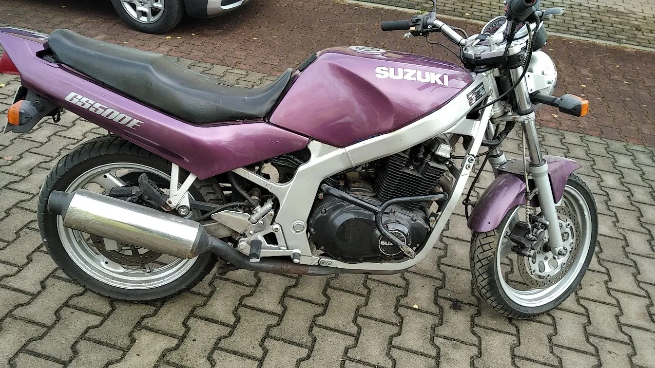 Fioletowy motocykl marki Suzuki, model GS500E, zaparkowany bokiem na kostce brukowej. Pojazd posiada srebrny wydech, czarne siedzenie i srebrne felgi.