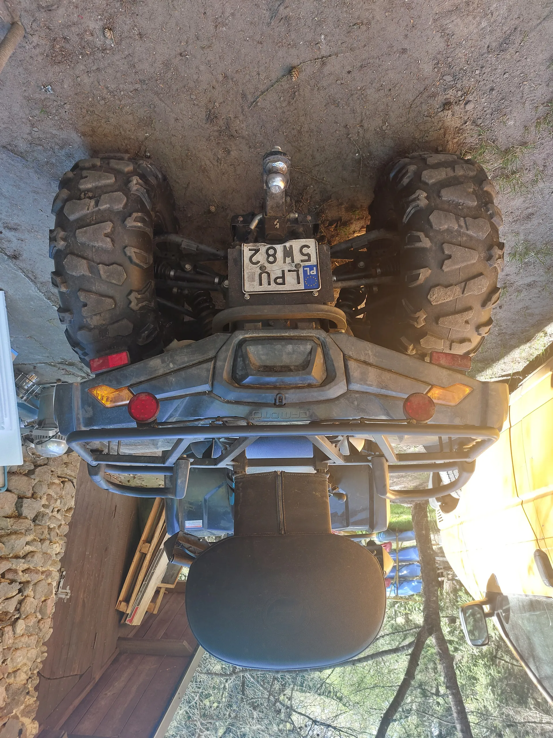 Czarny quad marki CFMOTO z pionowo umieszczoną tablicą rejestracyjną PL LPU 5M82. Quad stoi na brudnej ziemi, w tle kamienica z drewnianymi drzwiami, deski i drzewa. Widoczne są duże opony z głębokim profilem, czerwone światła tylna i żółte kierunkowskazy. Po lewej stronie obrazu fragment żółtego pojazdu.