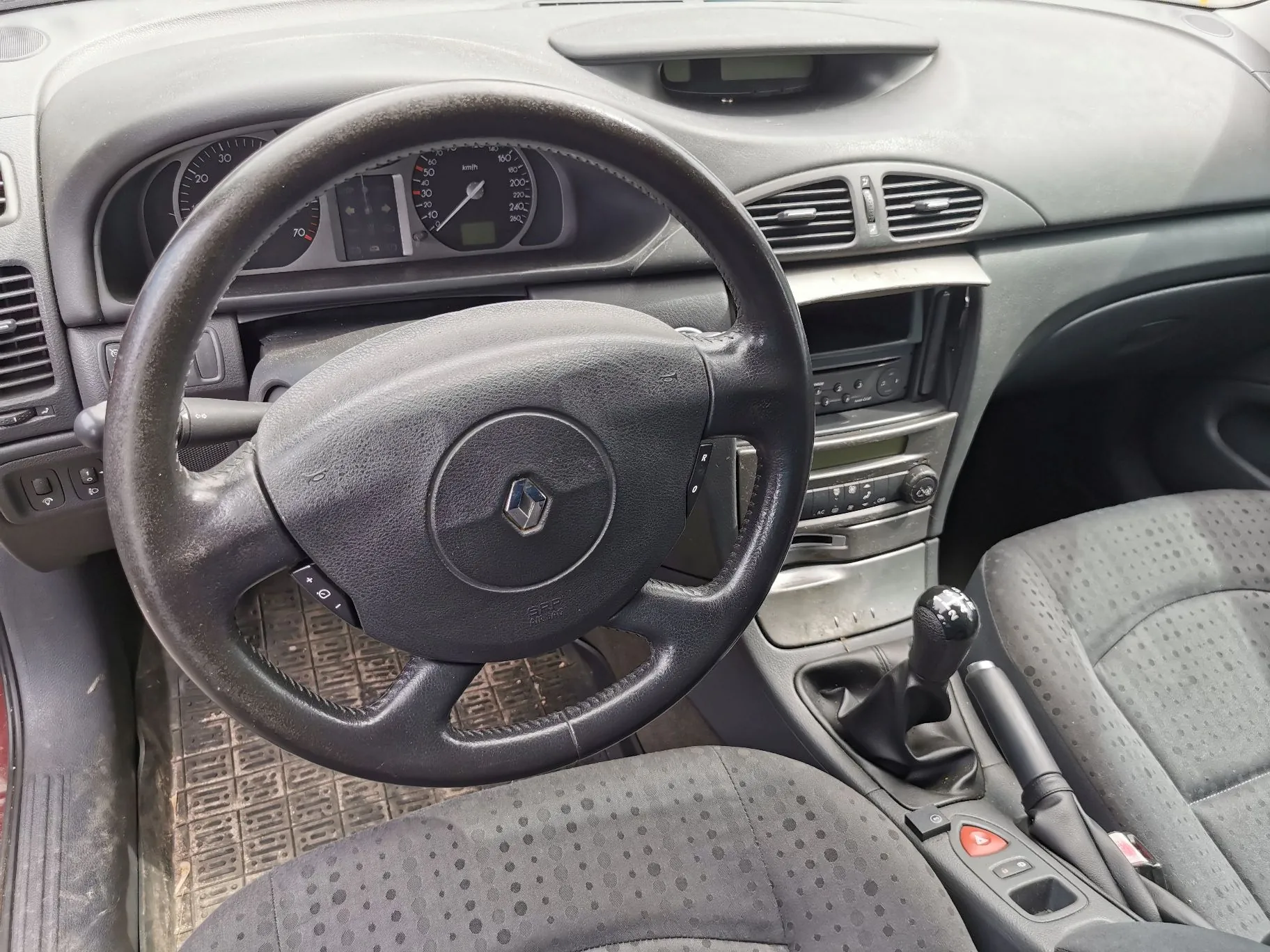 Wnętrze samochodu z perspektywy kierowcy. Widoczna czarna, skórzana kierownica z logo marki Renault w centrum. W tle znajduje się deska rozdzielcza z analogowymi wskaźnikami prędkości i obrotów, nawiewami oraz konsolą środkową z radiem i lewarkiem zmiany biegów. W dolnej części zdjęcia widoczny jest fragment ciemnoszarego, materiałowego fotela z kropkowanym wzorem oraz gumowa mata na podłodze.