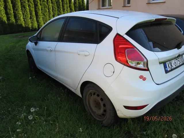 Biały samochód osobowy typu hatchback zaparkowany na trawie. Widoczna tylna część pojazdu z tablicą rejestracyjną KK-6750U oraz czerwonym naklejką w kształcie ust. Samochód znajduje się przed domem o beżowej elewacji z oknami. W tle wysokie zielone krzewy. Na trawie rosną białe kwiatki. W prawym dolnym rogu obrazu widoczna data 19/05/2026 pomarańczowym kolorem.