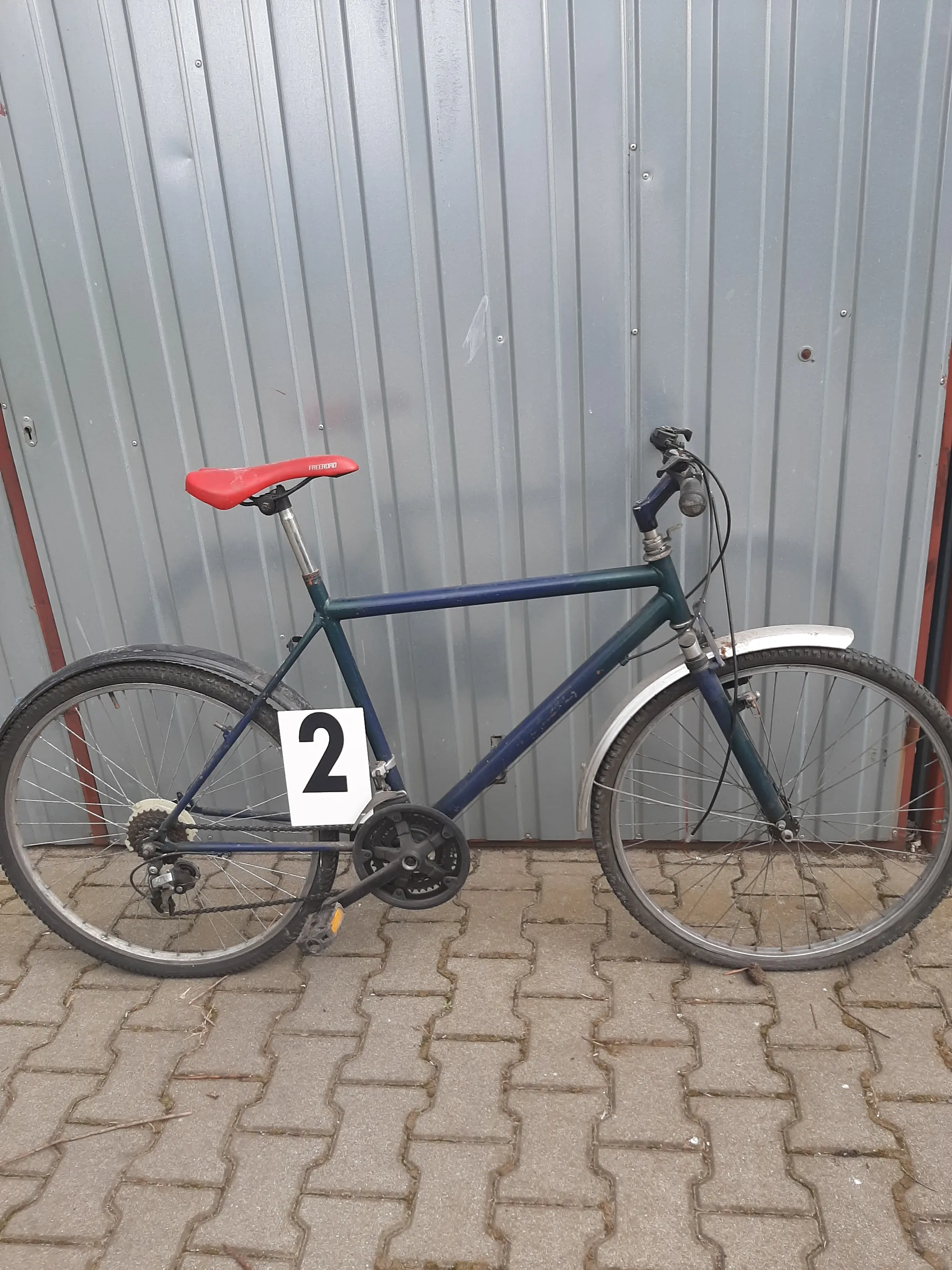 Niebieski rower z czerwonym siedziskiem oparty o szarą metalową bramę garażową. Na ramie roweru umocowana biała tabliczka z czarną cyfrą 2. Tło stanowi brukowana powierzchnia z kamiennymi płytami.