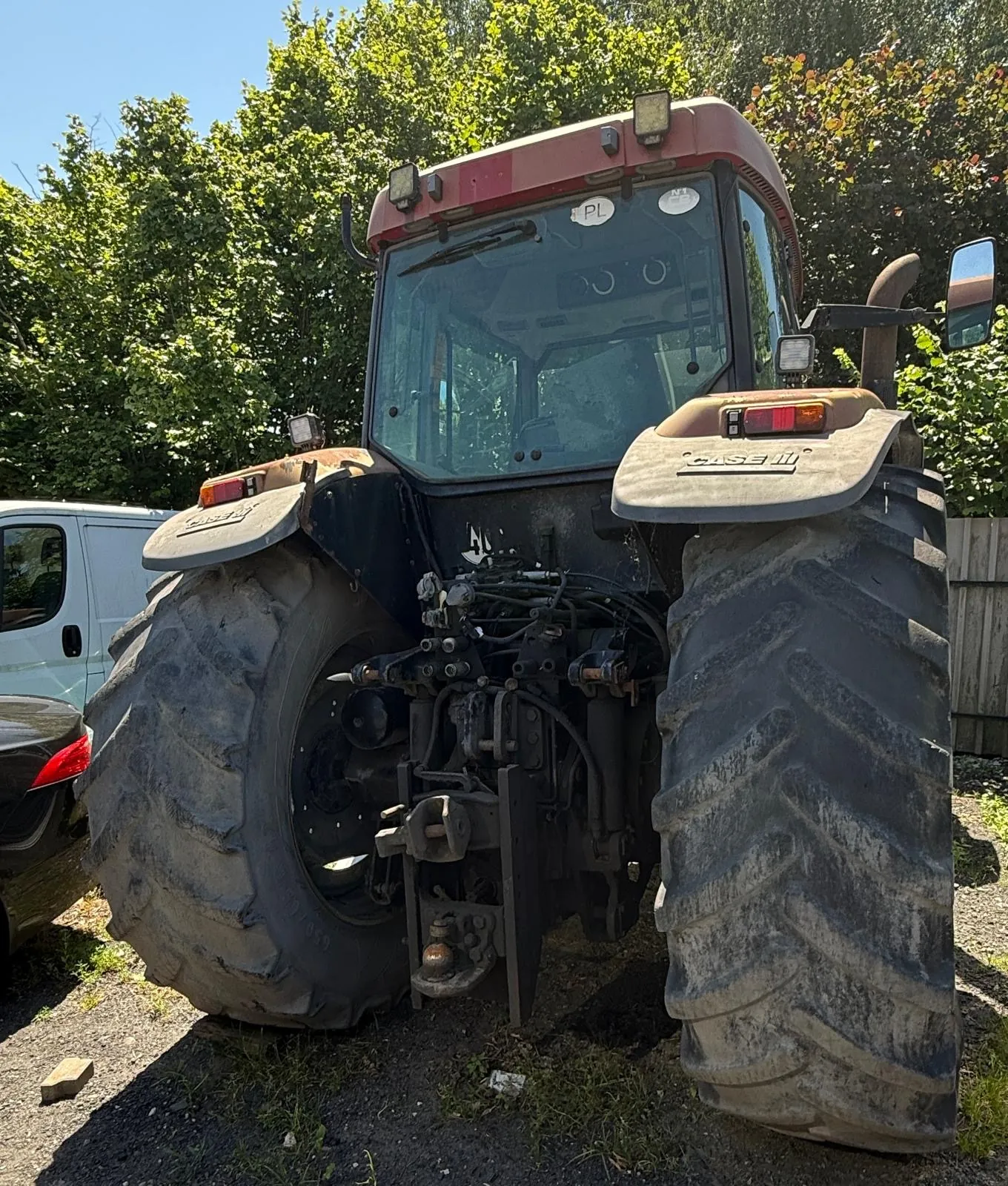 Widok z tyłu na czerwony traktor marki Case IH z dużymi, brudnymi oponami z wyraźnym bieżnikiem. Traktor posiada szklaną kabinę i systemy hydrauliczne z tyłu. W tle widoczne są zielone drzewa oraz fragment białego vana i czarnego samochodu po lewej stronie.