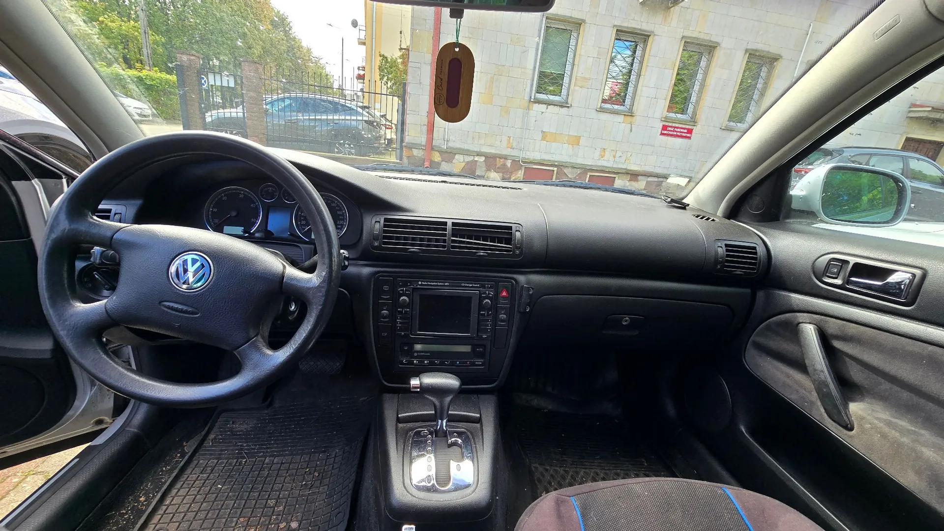 Widok z wnętrza samochodu marki Volkswagen, ukazujący deskę rozdzielczą, kierownicę z logo marki, zegary, radio z ekranem oraz lewarek zmiany biegów. Przez przednią szybę widoczny jest budynek z oknami oraz zaparkowane samochody.