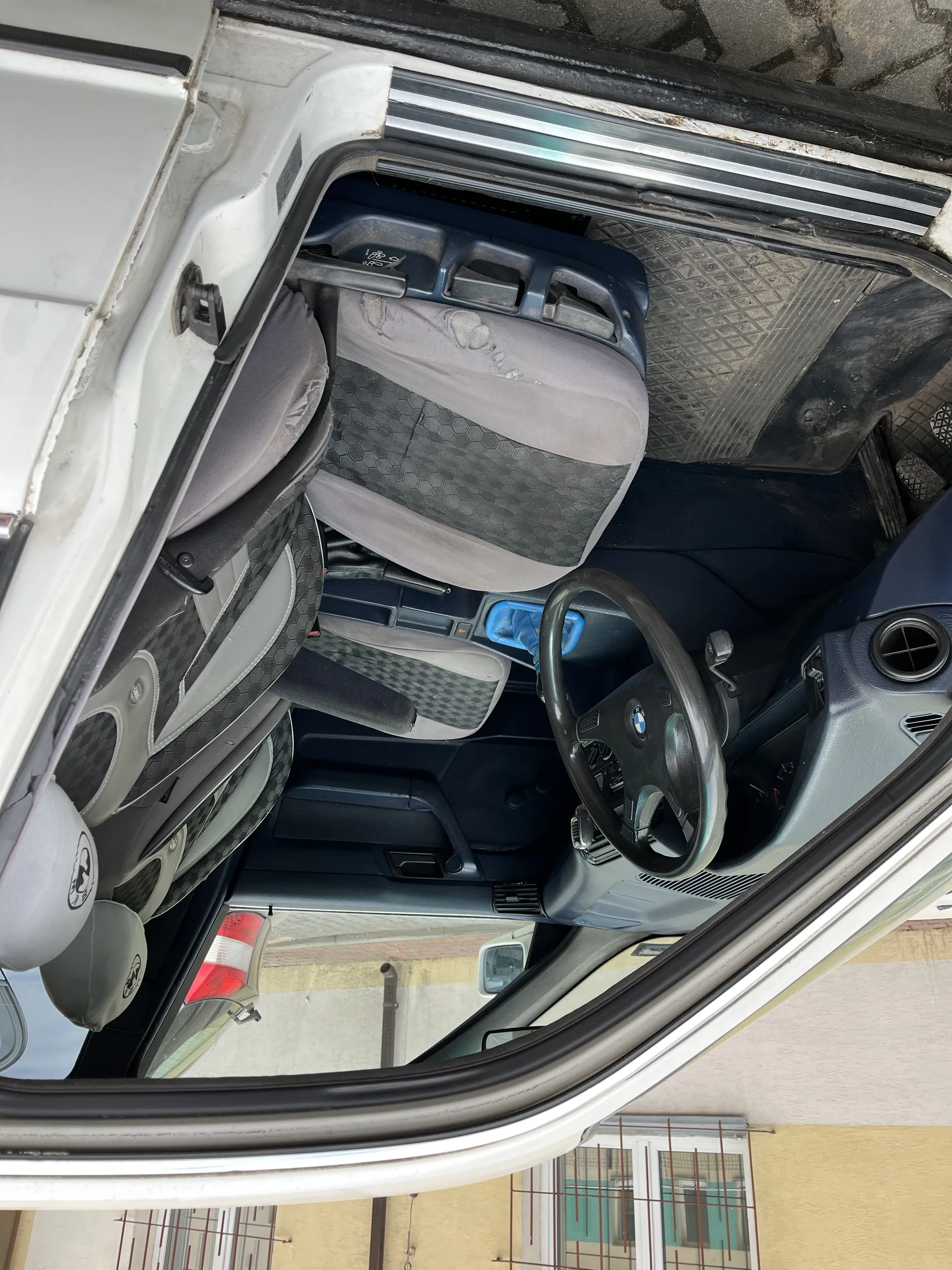 Wnętrze białego samochodu z otwartą drzwiami kierowcy. Widoczna kierownica z logo BMW, szare siedzenia z geometrycznym wzorem, jedno z siedzeń ma uszkodzenie. W centralnej konsoli znajduje się niebieski przedmiot. W tle budynek o żółtej elewacji z oknami i balustradami. Samochód zaparkowany na brukowanej powierzchni.