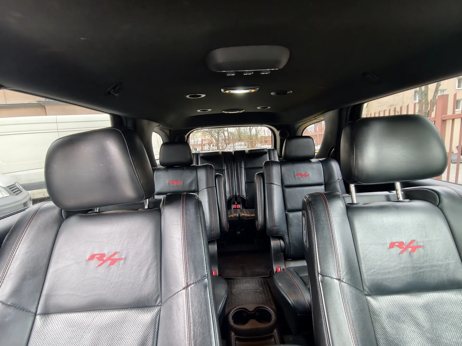 Zdjęcie przedstawia wnętrze samochodu z czarnymi skórzonymi siedzeniami z czerwonymi napisami "R/T" na oparciach. Widać kilka rzędów siedzeń, sufit z oświetleniem i otworem na dachu. Przez przednią szybę widoczne są budynki i ogrodzenie. Siedzenia mają czerwone przeszycia, a podłoga posiada czarny dywanik z uchwytem na kubki.