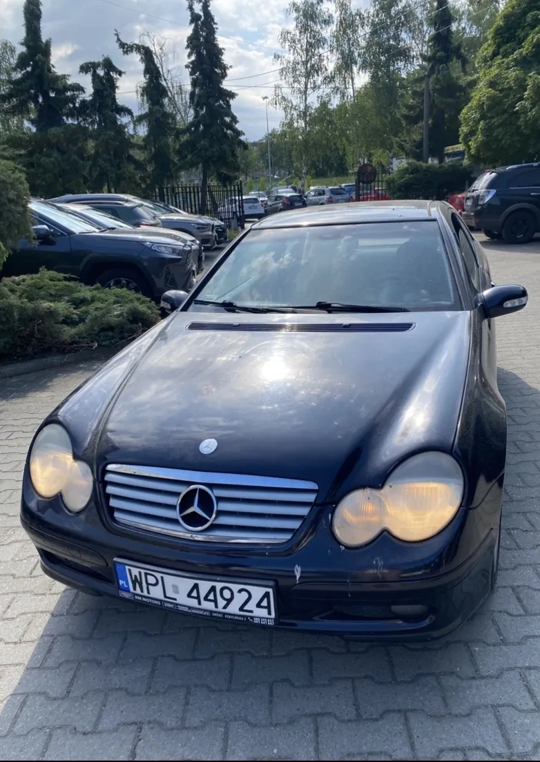 Czarny samochód marki Mercedes-Benz zaparkowany na brukowanym placu. Widoczna tablica rejestracyjna PL WPL 44924. W tle inne zaparkowane pojazdy, drzewa, ogrodzenie oraz częściowo zachmurzone niebo. Światła przednie samochodu są włączone, a na masce widoczny jest emblemat trójgwiazdy.