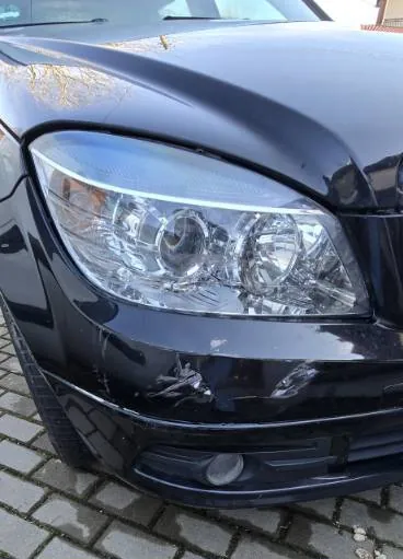 Czarny samochód z widocznym przodem, w tym reflektorem i zarysowanym zderzakiem z widocznymi uszkodzeniami. Samochód stoi na brukowanej powierzchni. W tle częściowo widoczne są drzewa i fragment budynku.