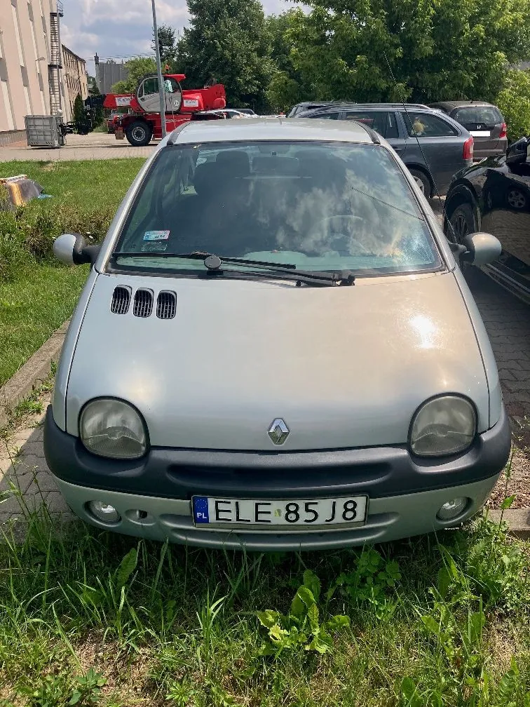 Srebrny samochód marki Renault zaparkowany na trawie. Widoczna jest przednia część pojazdu z tablicą rejestracyjną o numerze ELE 85J8. W tle znajdują się inne zaparkowane samochody, czerwony pojazd specjalistyczny oraz budynek i drzewa.