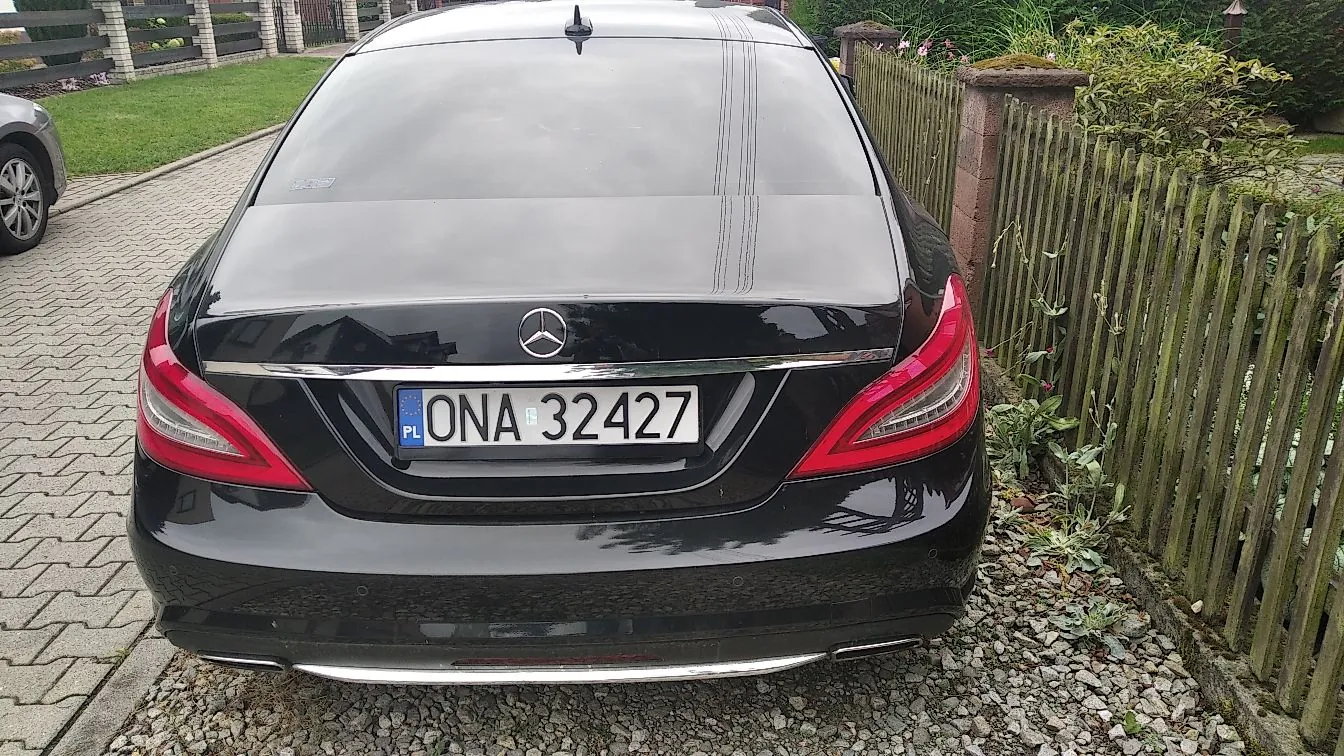 Tylny widok czarnego samochodu marki Mercedes-Benz zaparkowanego na podjeździe z kostki brukowej obok drewnianego ogrodzenia. Samochód posiada polską tablicę rejestracyjną o numerze ONA 32427.