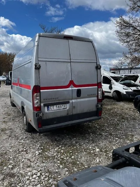 Srebrny furgon Peugeot z czerwoną linią na tylnych drzwiach, stojący na żwirowym placu. Widoczny tabliczka rejestracyjna CT 1661V. W tle białe furgony, samochód z otwartą maską, drzewa, budynek oraz niebo z chmurami.