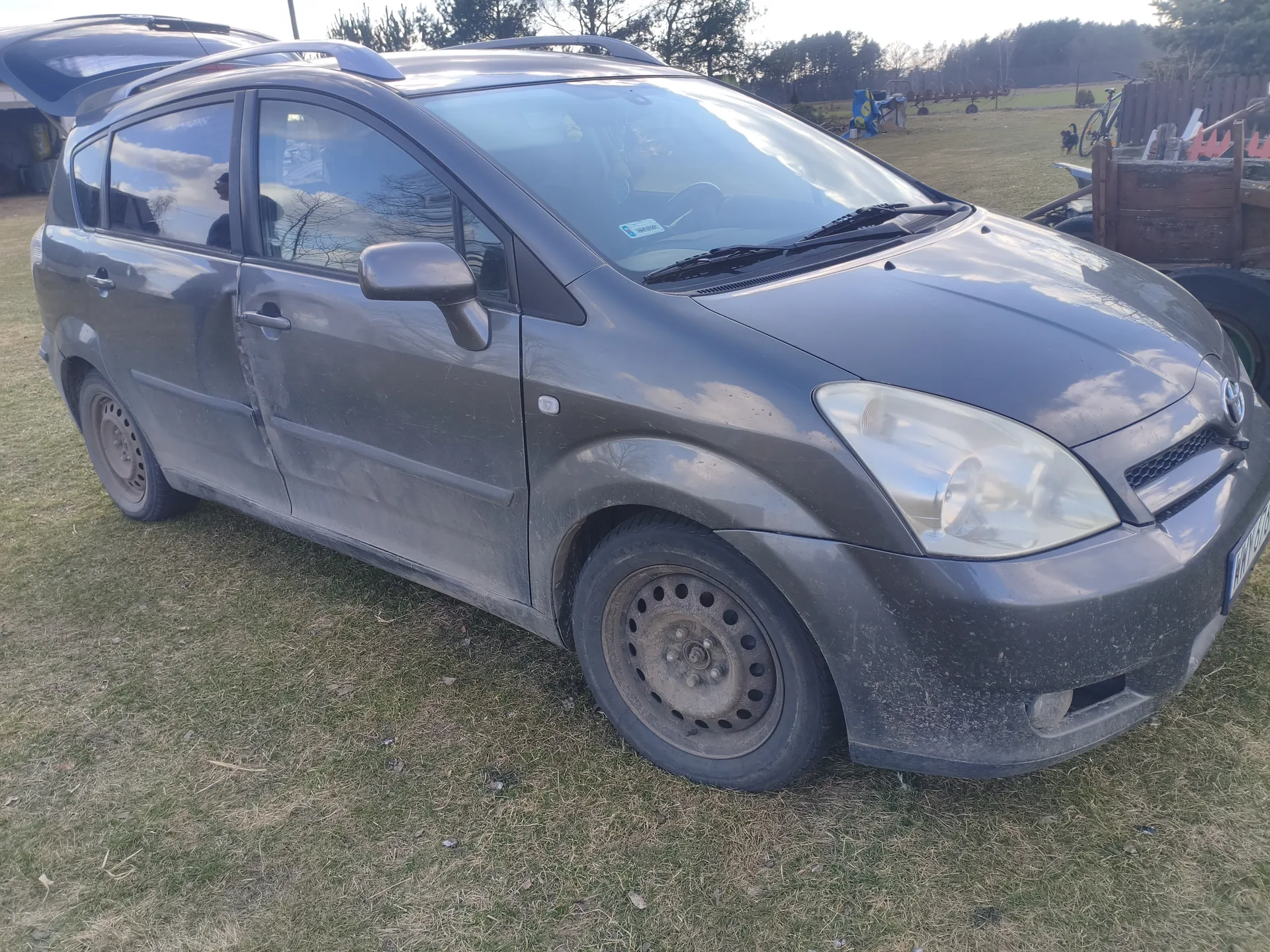 Szary samochód osobowy typu kombi z otwartym bagażnikiem i dachowym bagażnikiem, zaparkowany na trawie. Pojazd jest zabrudzony, widoczne są felgi bez opon na przednim kole. W tle polana, drzewa, rower, drewniana przyczepa oraz rolnicze narzędzia. Na przedniej szybie naklejka, tablica rejestracyjna częściowo widoczna po prawej stronie. Niebo częściowo zachmurzone.