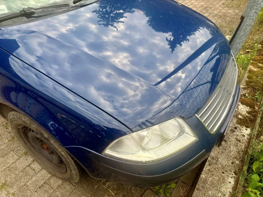 Zbliżenie na przód niebieskiego samochodu. Widoczna jest maska z odbiciem chmur i drzew, prawy reflektor oraz fragment srebrnego grilla. Samochód stoi na kostce brukowej obok metalowego słupka i zielonej roślinności.