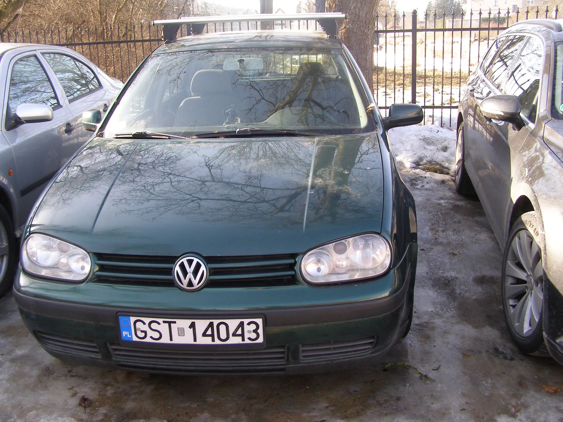 Zielony samochód marki Volkswagen Golf z tablicą rejestracyjną PL GST 14043, zaparkowany na zimowym placu parkingowym. Po obu stronach widoczne są inne pojazdy: srebrny samochód po lewej i szary po prawej. Na tle czarny metalowy płot, drzewa bez liści oraz śnieg na ziemi. Na dachu Volkswagena znajduje się biały dachówka. Odbicia gałęzi drzew na przedniej szybie i kapotzie.