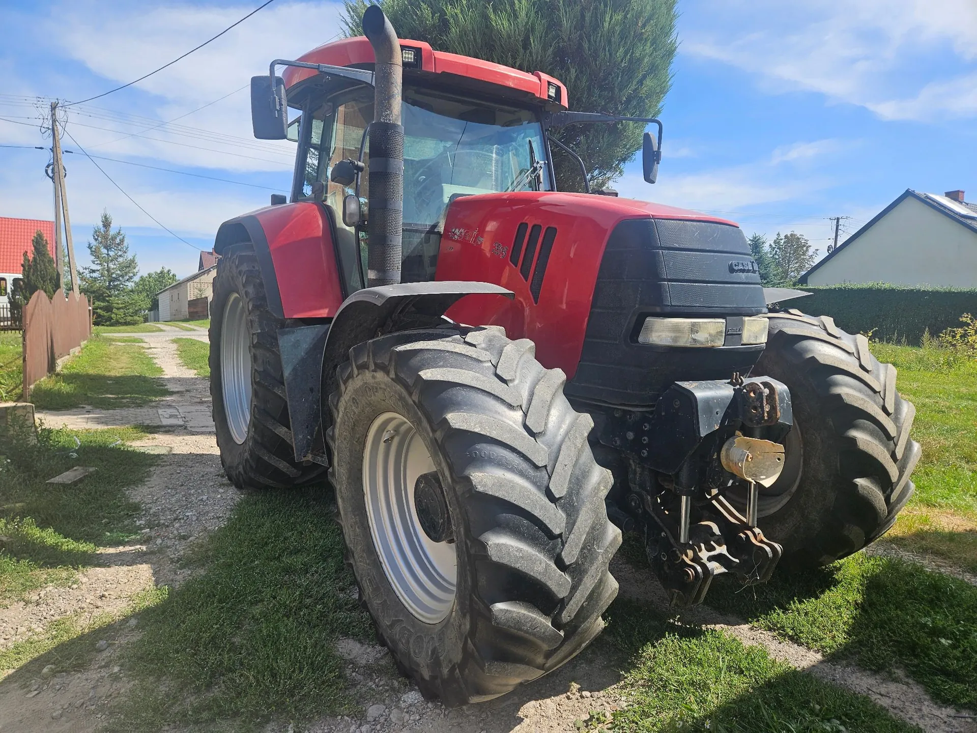 Czerwony traktor marki Case IH zaparkowany na piaszczystej drodze w wiejskiej okolicy. Maszyna posiada duże, czarne opony z wyraźnym bieżnikiem, wysoką kabinę z przeszklonymi szybami oraz pionową rurę wydechową. W tle widoczne są zielone drzewa, krzewy, fragmenty domów oraz niebieskie niebo z lekkimi chmurami.