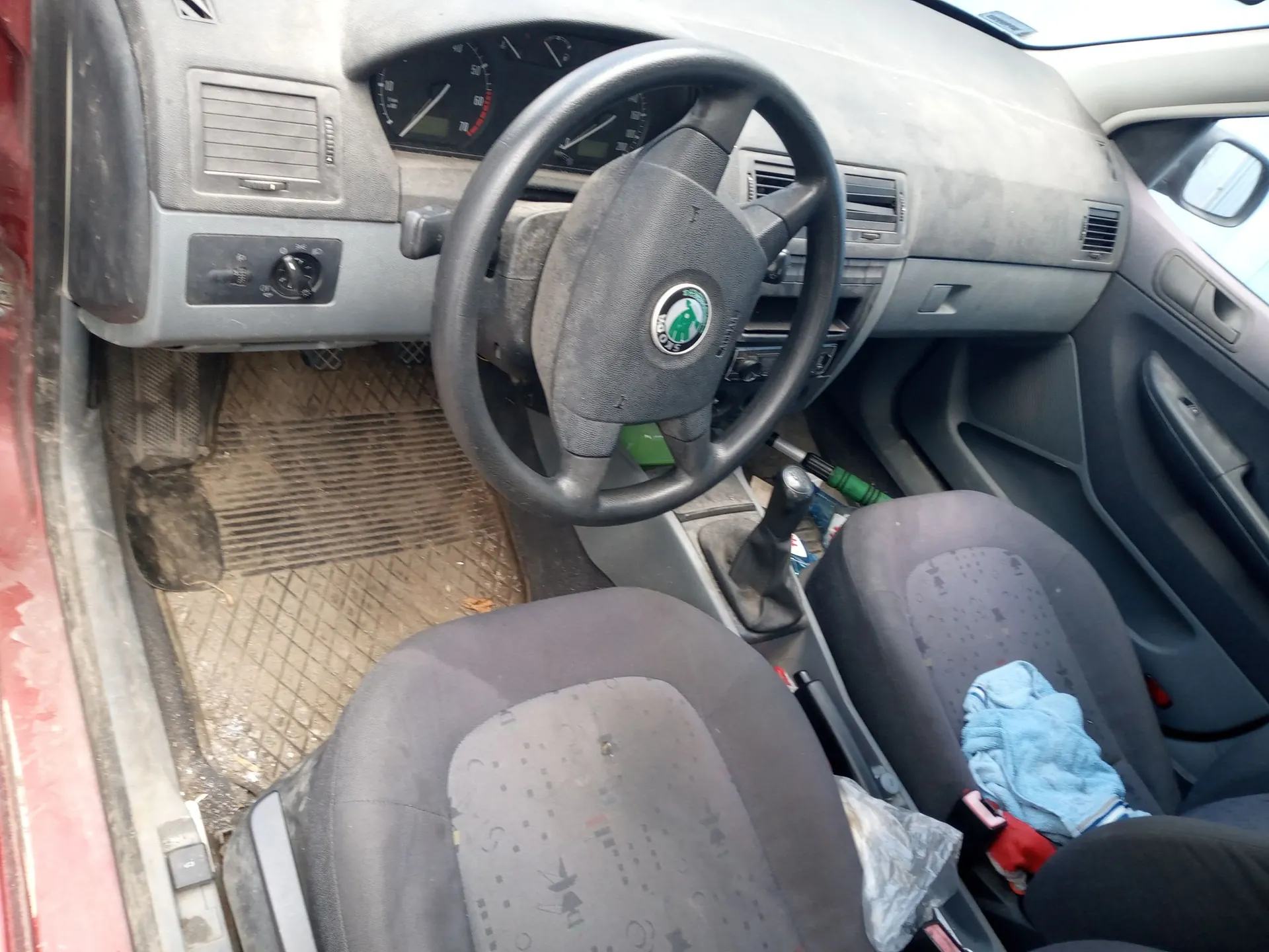 Wnętrze samochodu od strony kierowcy. Widoczna jest zakurzona deska rozdzielcza, kierownica z logo marki Skoda, fotele z ciemnoszarej tkaniny oraz gumowe dywaniki na podłodze. Wnętrze jest zaniedbane i pokryte warstwą kurzu.