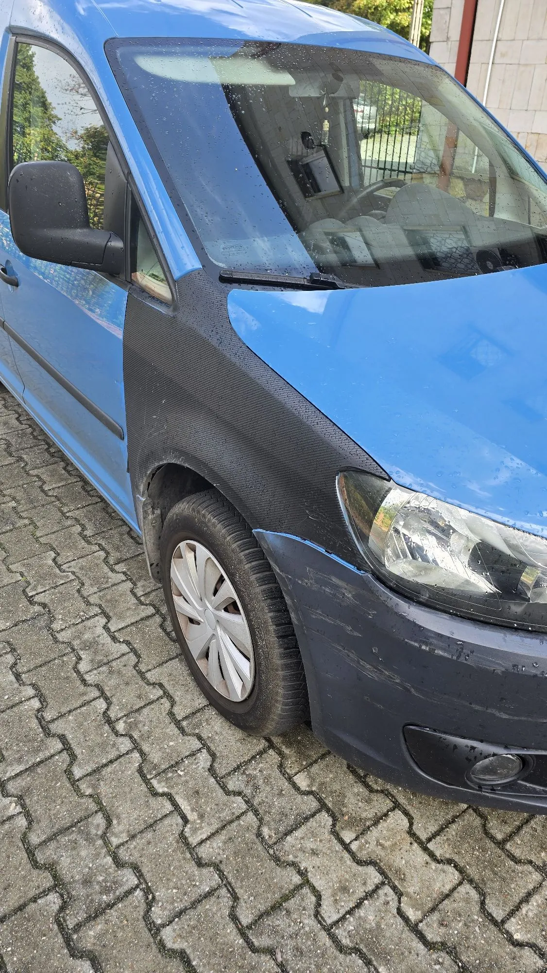 Zbliżenie na przód niebieskiego samochodu typu van zaparkowanego na kostce brukowej. Przedni lewy błotnik jest zastąpiony czarnym elementem wykonanym z włókna węglowego lub materiału go imitującego. Widoczna jest przednia lampa oraz koło z plastikową osłoną.