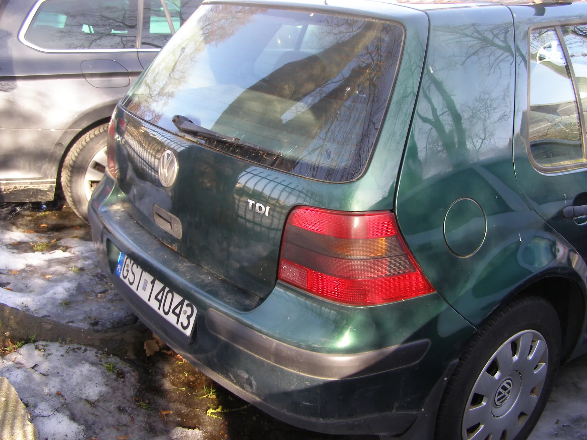 Zielony samochód Volkswagen typu hatchback widziany od tyłu. Na bagażniku widoczny emblemat marki oraz napis 'TDI'. Tablica rejestracyjna PL GST 14043. W tle częściowo widoczny szary samochód. Na ziemi śnieg i plamy trawy.