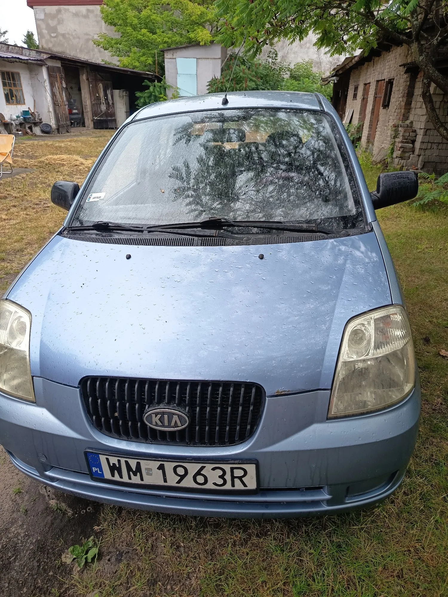 Jasnoniebieski samochód marki Kia zaparkowany na trawie przed starymi budynkami. Auto jest mokre od deszczu. Na przodzie widoczny jest czarny grill z logo KIA oraz tablica rejestracyjna o numerze WM 1963R z oznaczeniem PL.