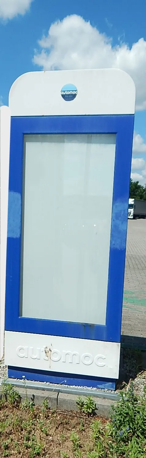 Pionowy, biało-niebieski słup informacyjny z pustą, przeszkloną witryną w centrum. Na górze znajduje się małe logo z napisem 'automoc POLSKA', a na dole duży, wypukły napis 'automoc'. Obiekt stoi na betonowej podstawie na zewnątrz, w tle widoczne jest niebo z chmurami oraz fragment białej ciężarówki.
