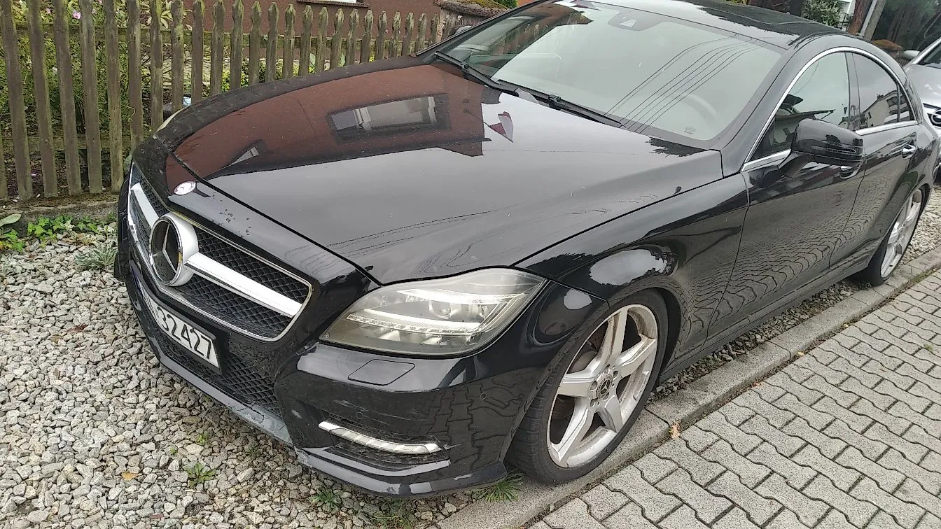 Czarny samochód marki Mercedes-Benz zaparkowany przy krawężniku na kostce brukowej. Widoczny jest przód i bok pojazdu z charakterystycznym logo gwiazdy na grillu oraz srebrnymi felgami. W tle znajduje się drewniany płot.