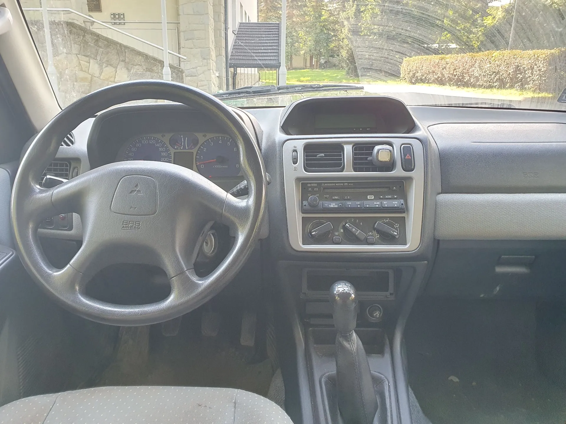 Widok z wnętrza samochodu na deskę rozdzielczą i kierownicę. Po lewej stronie znajduje się czarna, czteroramienna kierownica z logo Mitsubishi w centrum i napisem 'SRS AIRBAG'. Za kierownicą widoczny jest panel wskaźników z prędkościomierzem i obrotomierzem. W centralnej części deski rozdzielczej znajdują się kratki nawiewu, radio z wyświetlaczem oraz pokrętła sterowania klimatyzacją. Poniżej znajduje się dźwignia zmiany biegów w czarnej obudowie. Przez przednią szybę widoczny jest fragment budynku z jasnego kamienia, płot oraz zielony żywopłot.