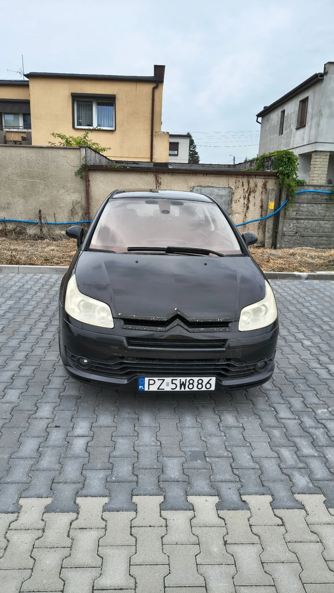Czarny samochód osobowy marki Citroën zaparkowany przodem na kostce brukowej. Pojazd ma polską tablicę rejestracyjną o numerze PZ 5W886. W tle widoczny jest jasny, dwupiętrowy budynek oraz betonowy mur z roślinnością.