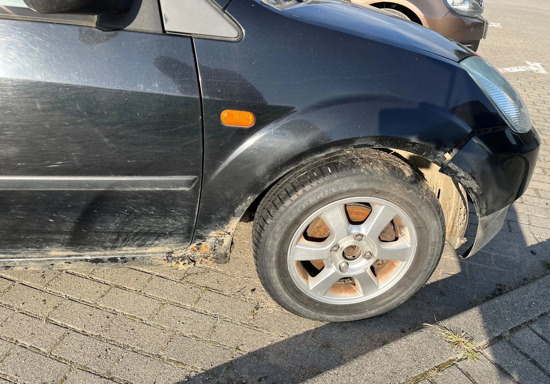 Zbliżenie na przednią prawą część czarnego samochodu. Widoczny jest przedni prawy nadkole z wyraźnymi śladami korozji i rdzy na dolnej krawędzi błotnika. Samochód posiada srebrną felgę aluminiową z pięcioma ramionami i oponę. Na boku nadkola znajduje się mały, pomarańczowy kierunkowskaz. Pojazd stoi na kostce brukowej.