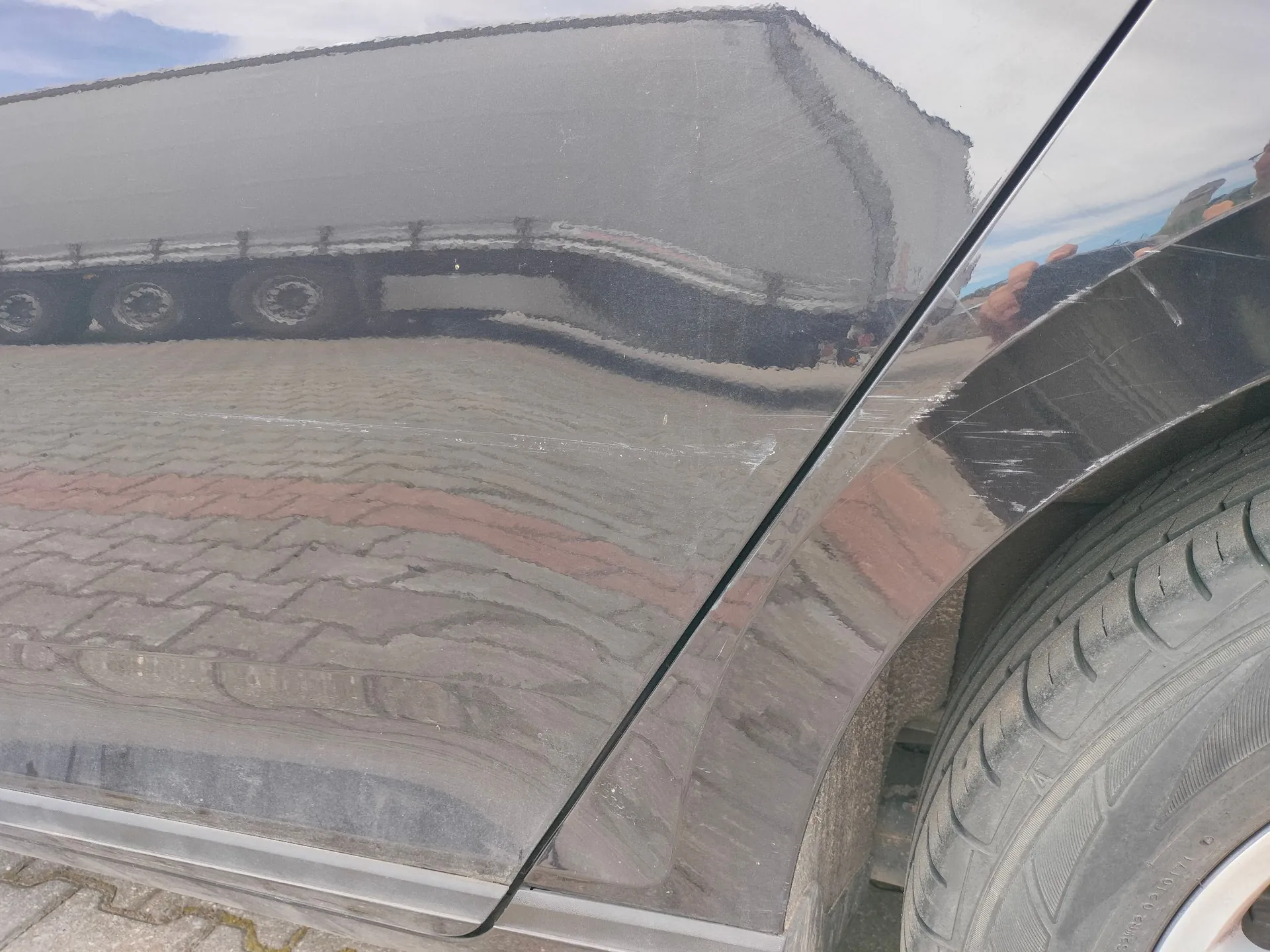 Zbliżenie na czarną, błyszczącą karoserię samochodu w okolicach tylnego nadkola. Na lakierze widoczne są liczne zarysowania oraz odbicie dużego, szarego samochodu ciężarowego z naczepą oraz brukowanej nawierzchni parkingu.