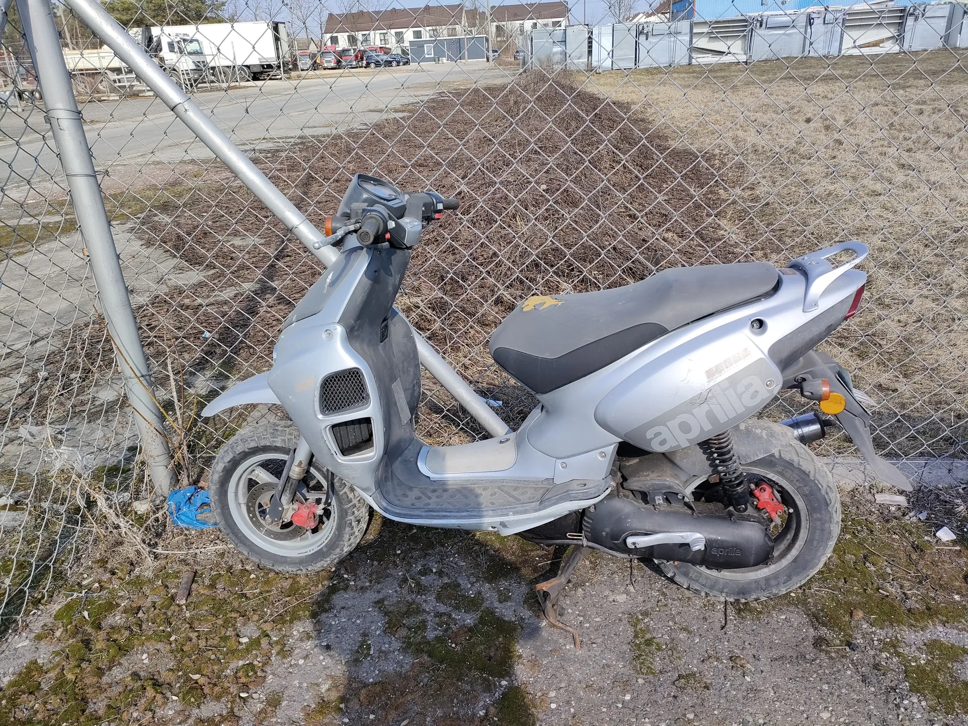 Srebrny skuter marki Aprilia zaparkowany przy siatce ogrodzeniowej. Skuter ma czarne siedzenie z żółtym motywem, widoczne są czerwone elementy hamulcowe i wydech. W tle siatka, sucha trawa, sterta ziemi, droga z pojazdami (biały naczepowy ciężarówka, inne samochody) oraz budynki przemysłowe. Na ziemi widoczne są plamy mchu i żwirek.