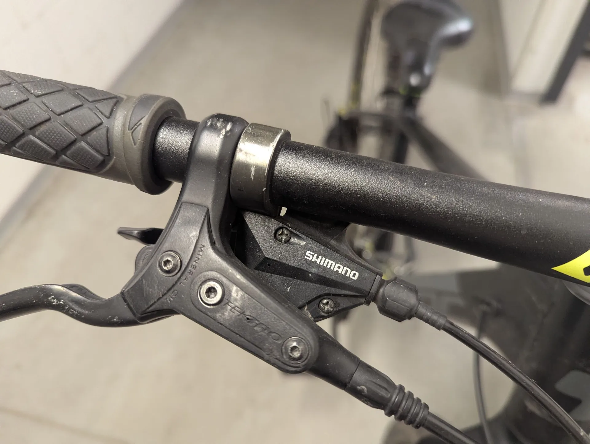 Zbliżenie na czarną klamkę hamulca hydraulicznego marki Shimano zamontowaną na kierownicy roweru. Na obudowie klamki widnieje napis 'SHIMANO' oraz mniejszy napis 'MINERAL OIL'. Widoczne są śruby mocujące oraz przewody hamulcowe. Kierownica jest czarna z gumową rączką o teksturowanej powierzchni.