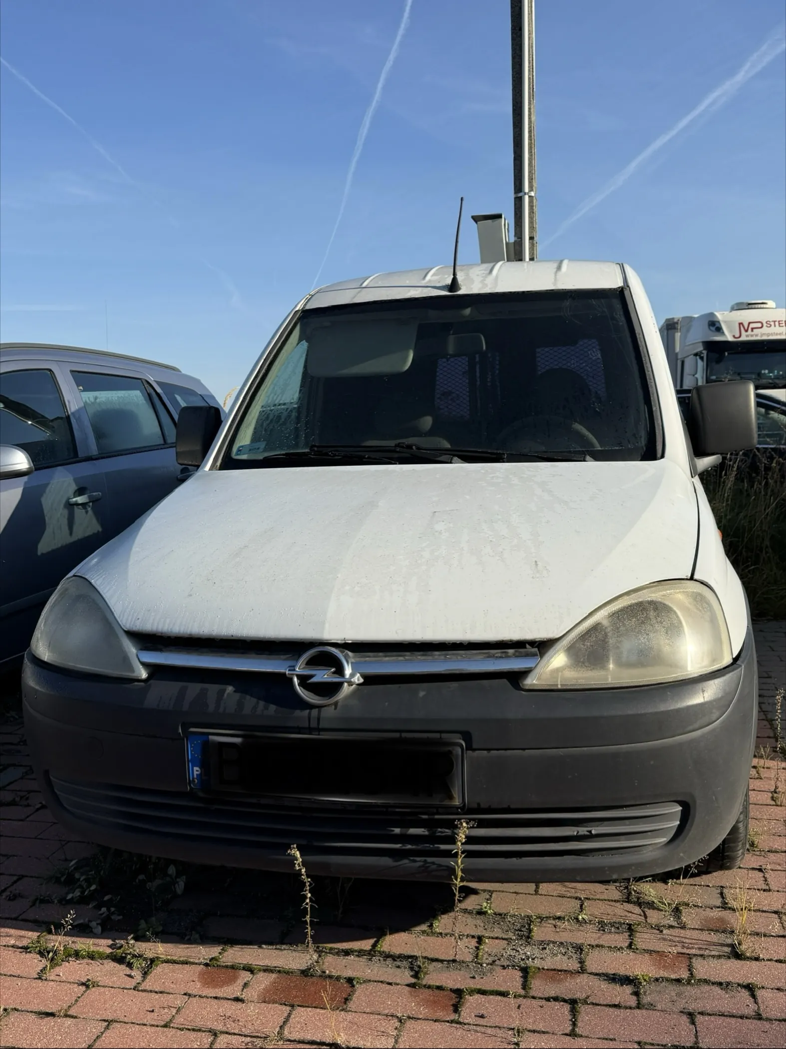 Biały samochód marki Opel Combo zaparkowany na kostce brukowej. Widoczny jest przód pojazdu z logo producenta na masce i zmatowiałymi reflektorami. Tablica rejestracyjna jest zakryta czarnym polem, widoczny jest jedynie niebieski pasek z literą B. W tle widać fragment innego srebrnego samochodu oraz białą ciężarówkę pod czystym, niebieskim niebem.
