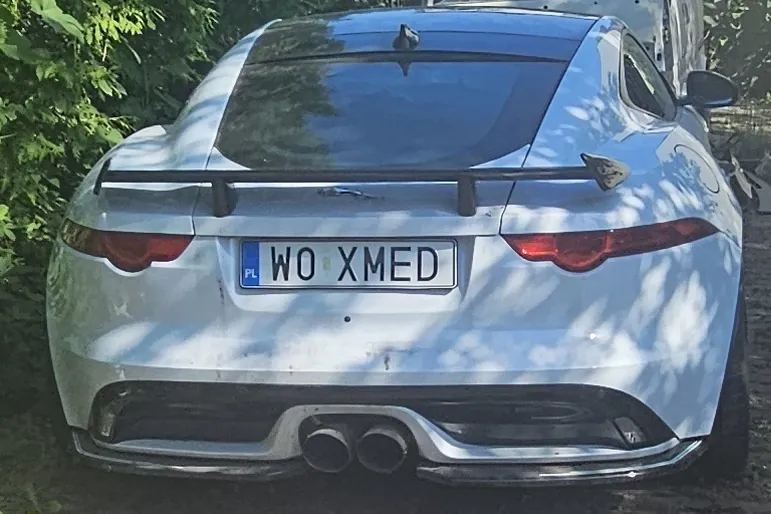Tylny widok srebrnego samochodu sportowego z czarnym spojlerem. Na samochodzie znajduje się polska tablica rejestracyjna o treści WO XMED. Auto zaparkowane jest na nieutwardzonym terenie w pobliżu zielonych krzewów.