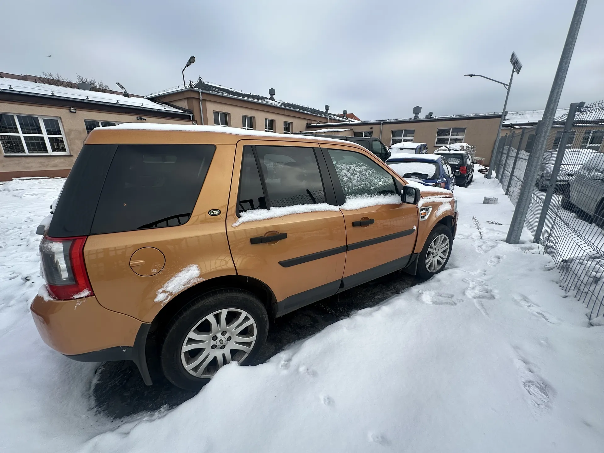 Złoty SUV zaparkowany na śnieżnym parkingu, pokryty śniegiem na dachu i szybach. W tle budynki o beżowych elewacjach z śniegiem na dachach, inne zaparkowane samochody oraz metalowe ogrodzenie. Na ziemi śnieg z widocznymi śladami stóp. Niebo zachmurzone.